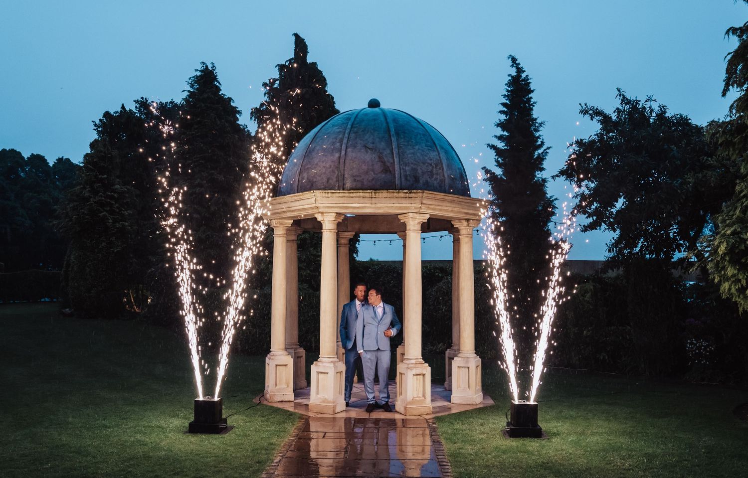 Couple stands in gazebo at dusk with dramatic sparkler fountains illuminating the scene.