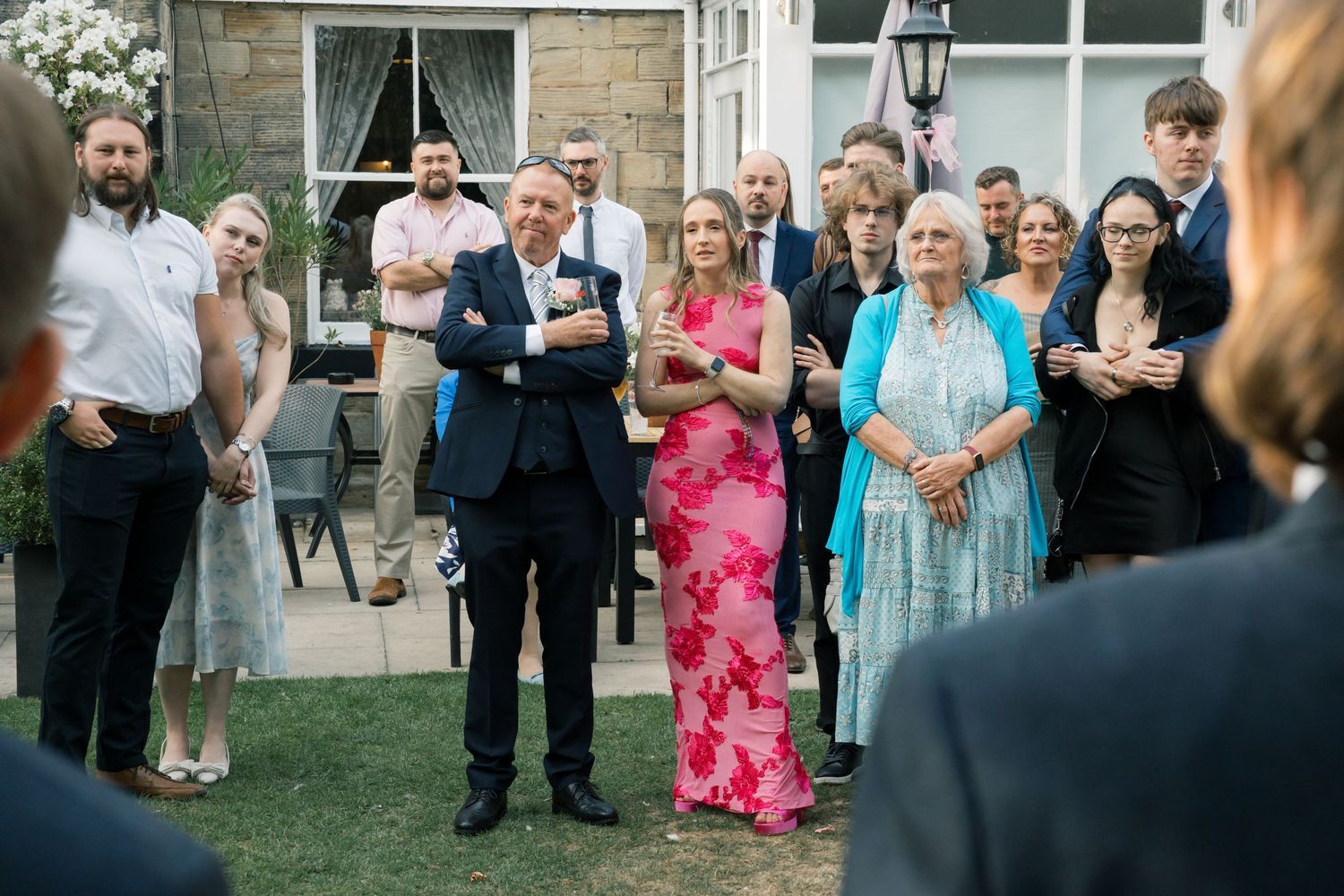 Wedding guests watching ceremony in garden setting with stone building in background.