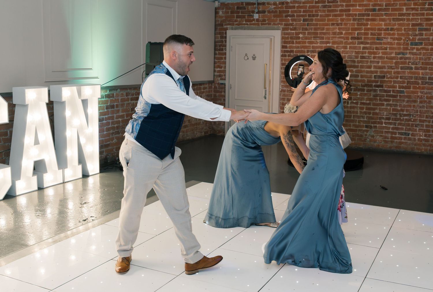 Wedding party dancing and celebrating on white dance floor with brick wall backdrop.