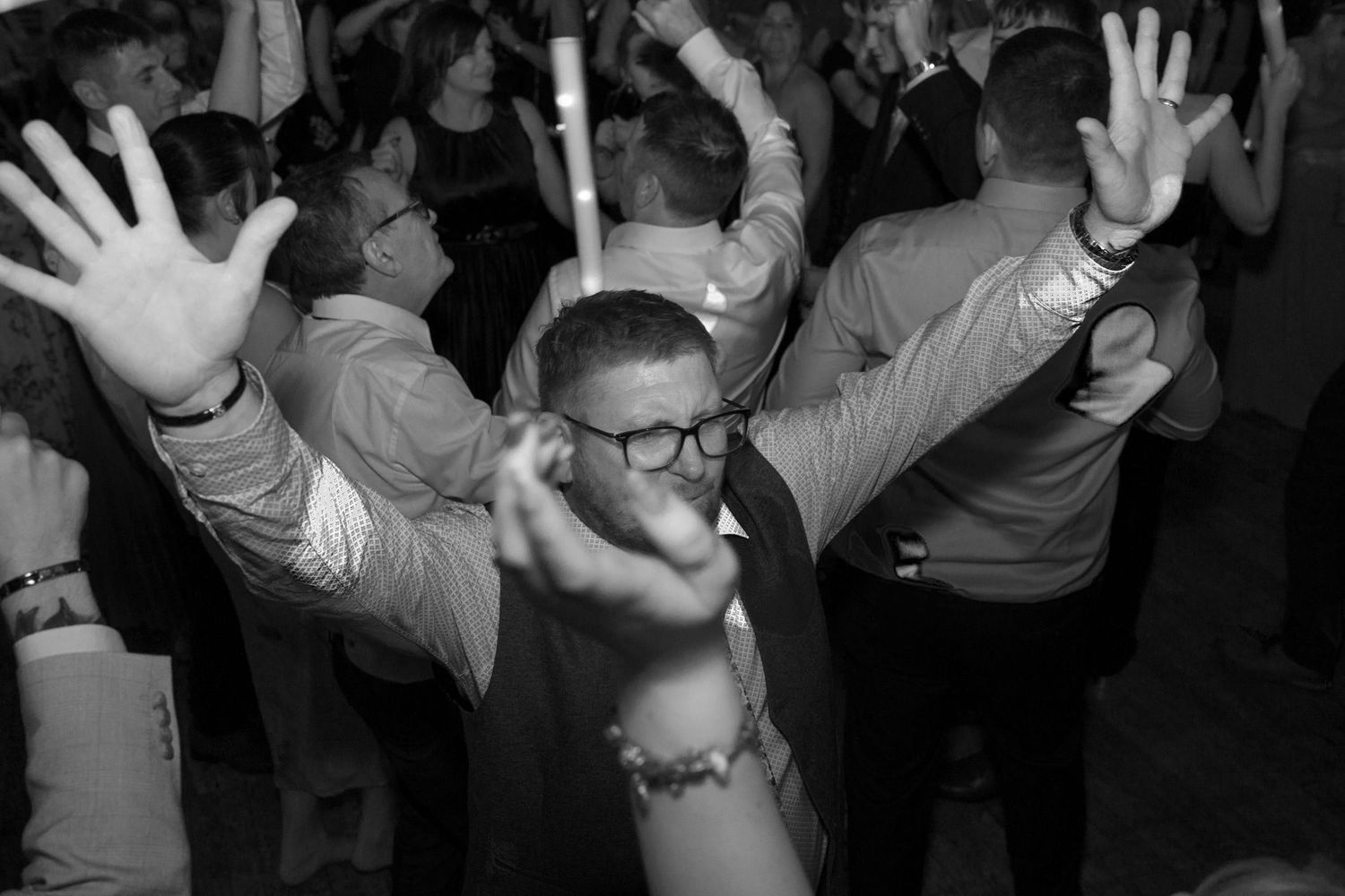 Wedding guests dancing with raised arms on a dimly lit dance floor.