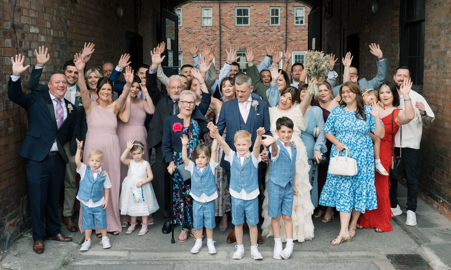 Large group wedding photo with guests in coordinated blue outfits posing against brick wall.