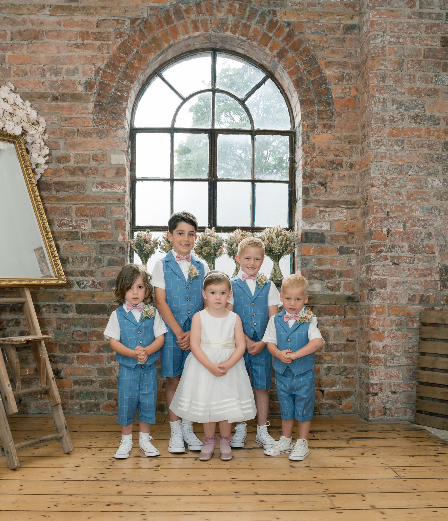 Young wedding party members pose in front of rustic brick wall with arched window.