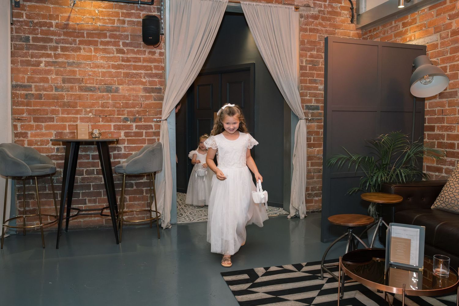 A young flower girl in a white dress twirls in front of exposed brick walls and white curtains at a wedding venue.