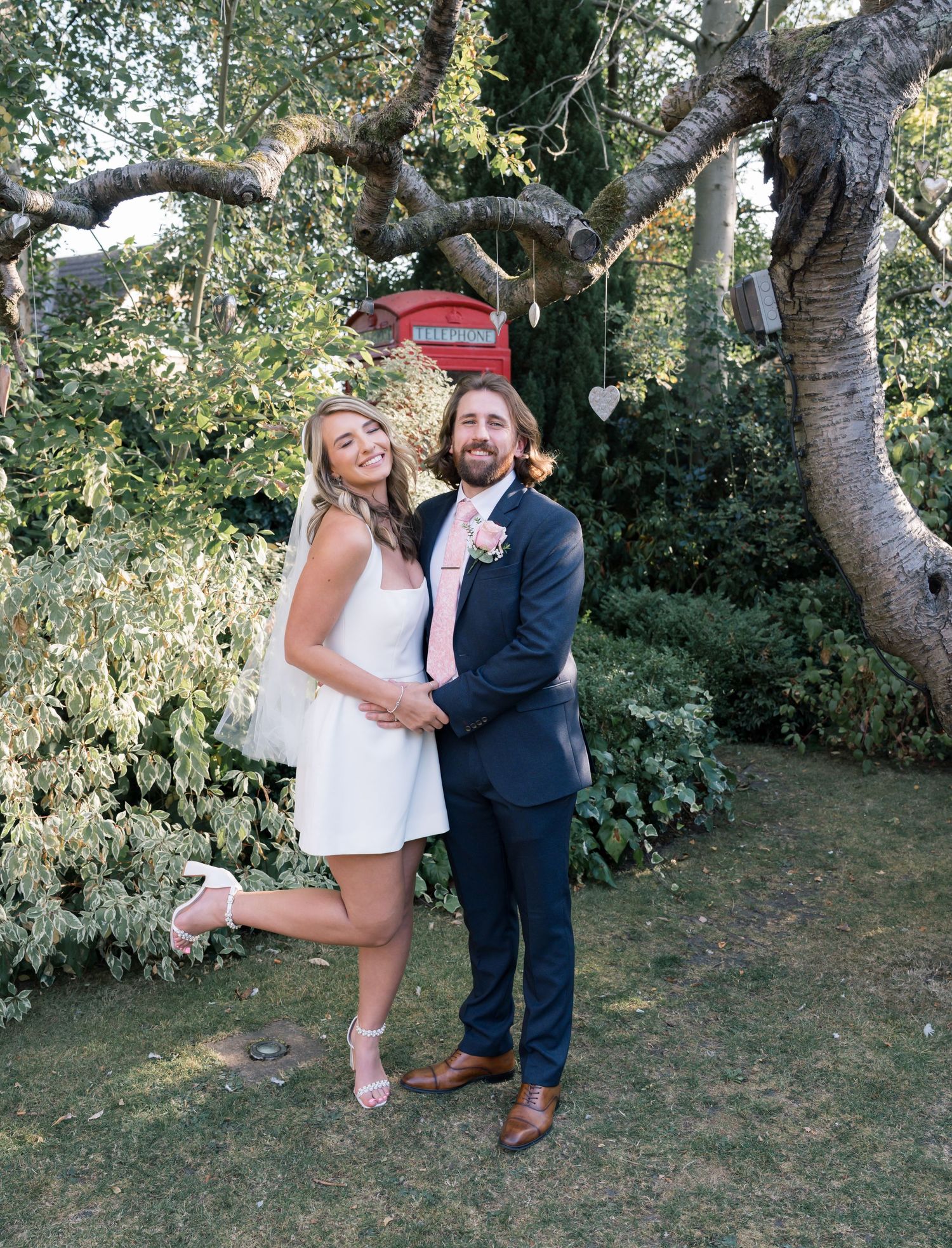 Wedding couple posing playfully under a large tree in an outdoor garden setting.