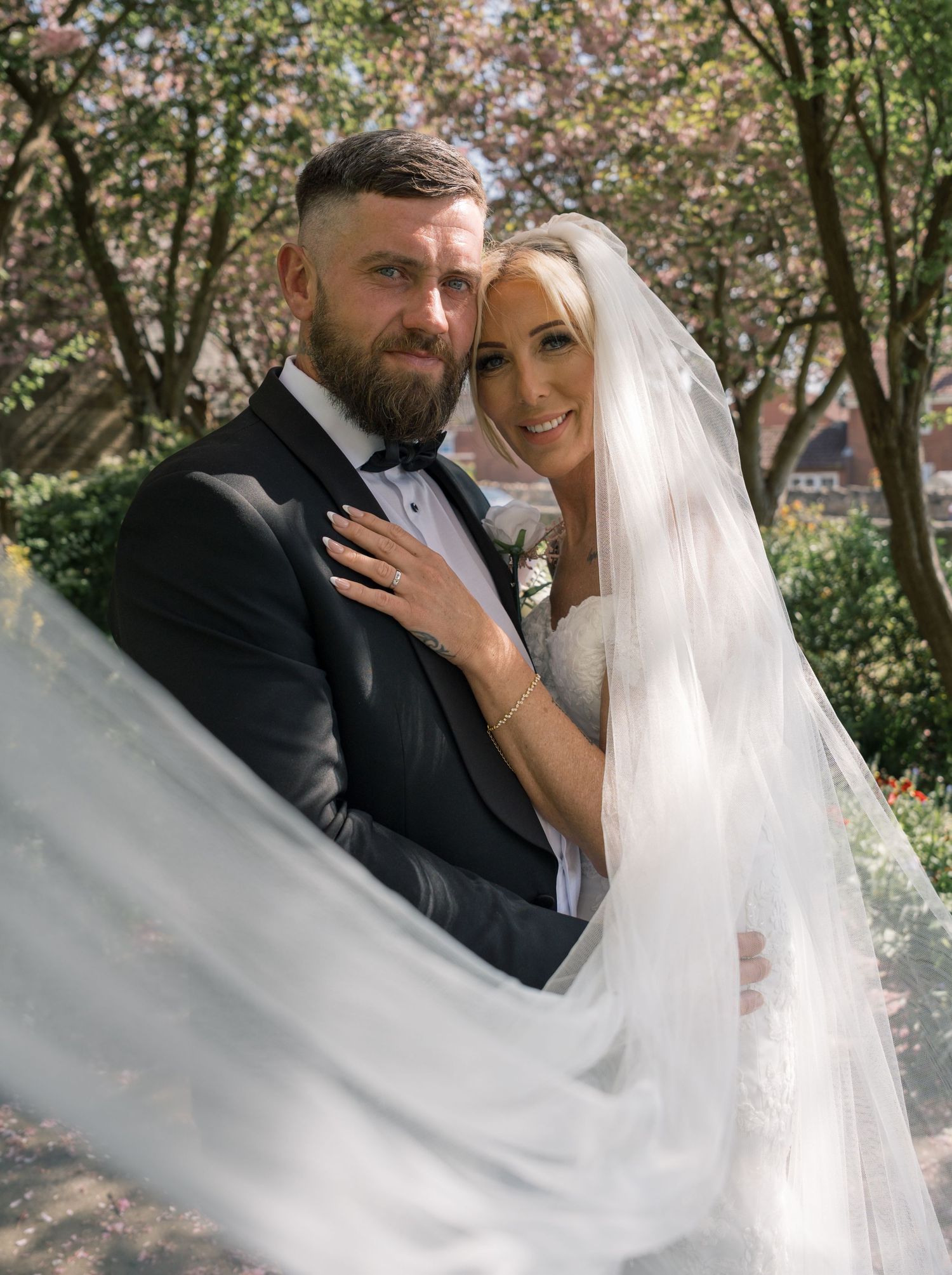 Bride in white gown and veil embraces groom in black tuxedo outdoors under blooming cherry blossom trees.