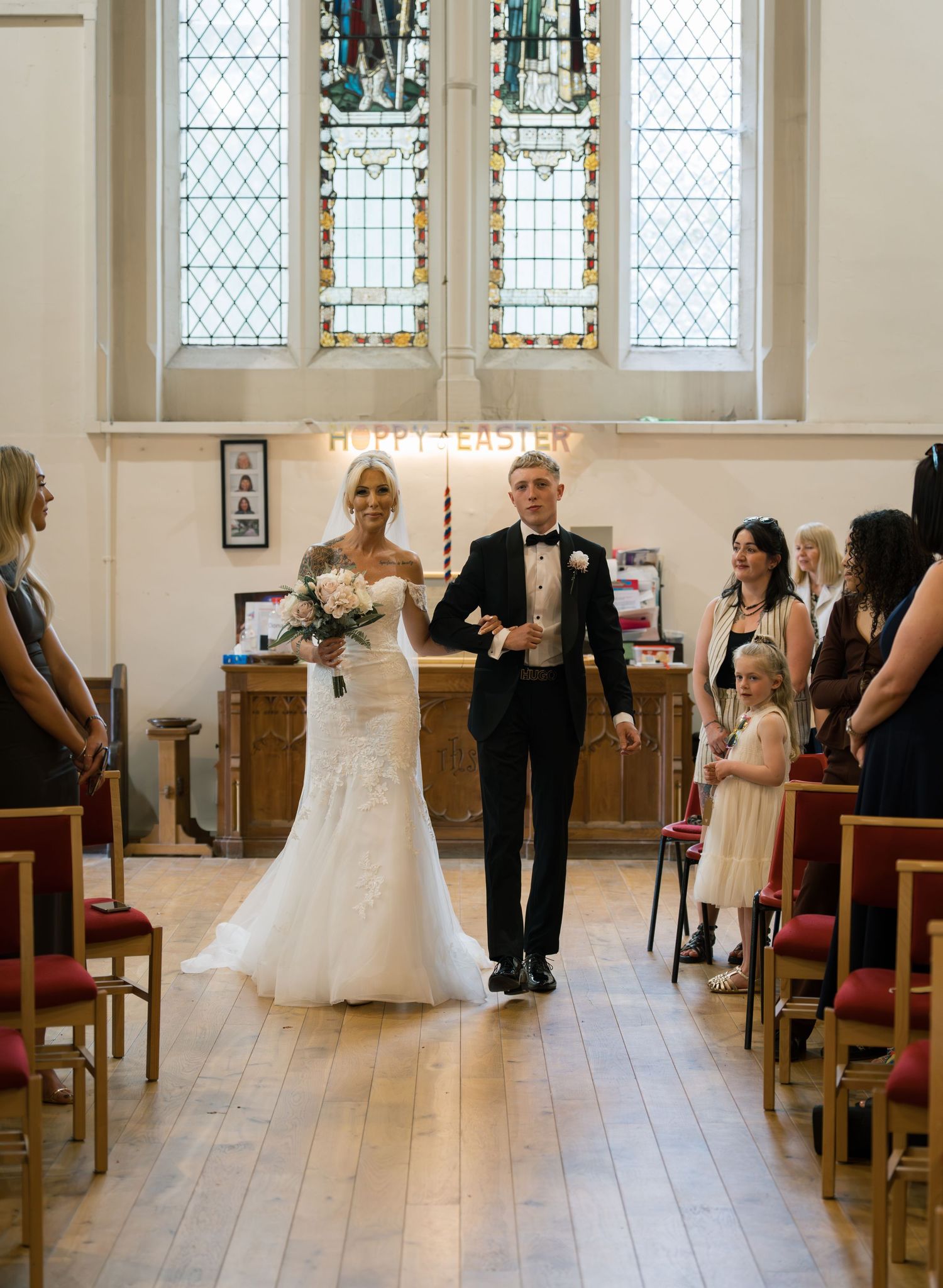 Bride and groom walking down the aisle of a church after their wedding ceremony, with guests watching and stained glass windows.