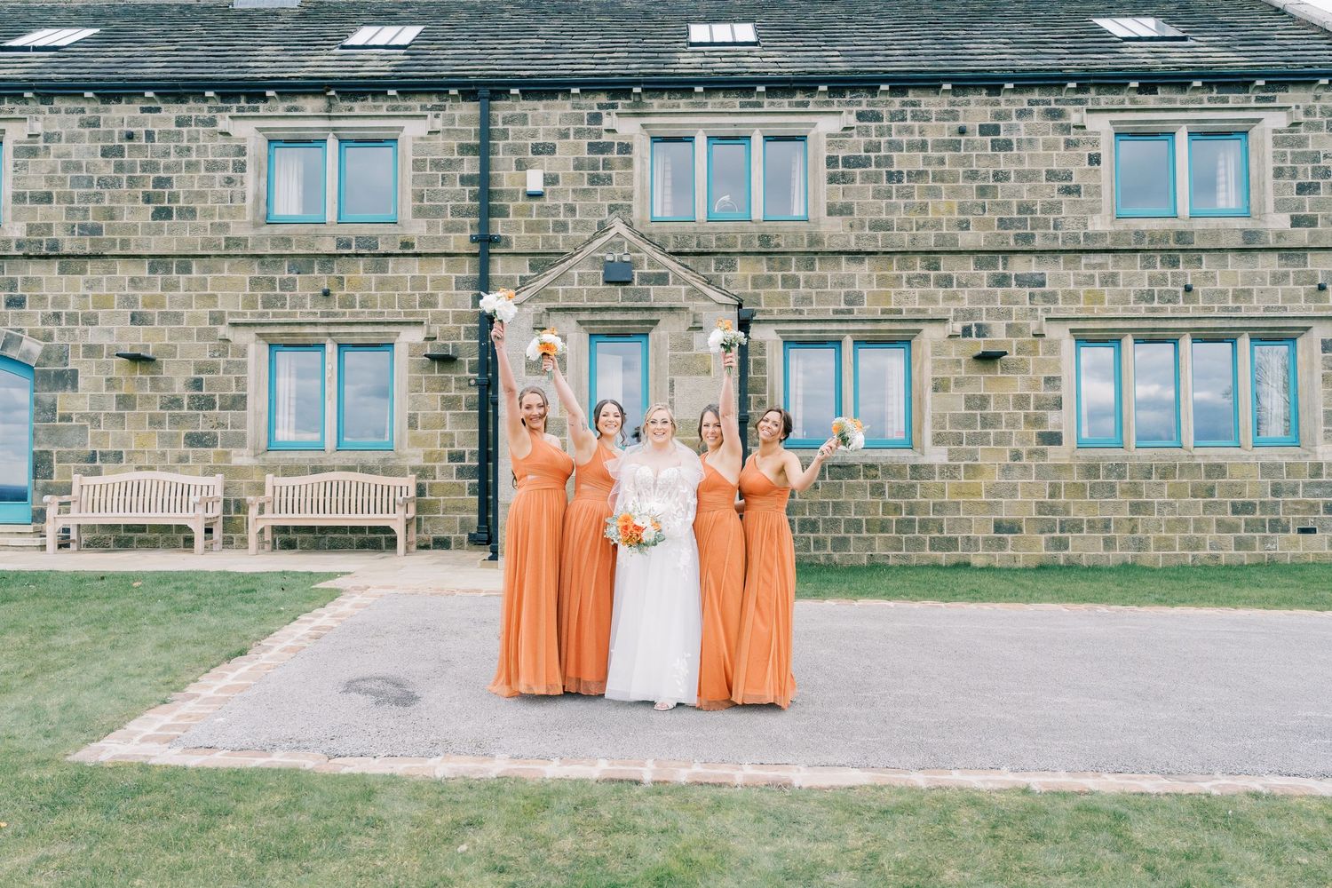 Wedding party in orange dresses poses in front of stone building with benches.