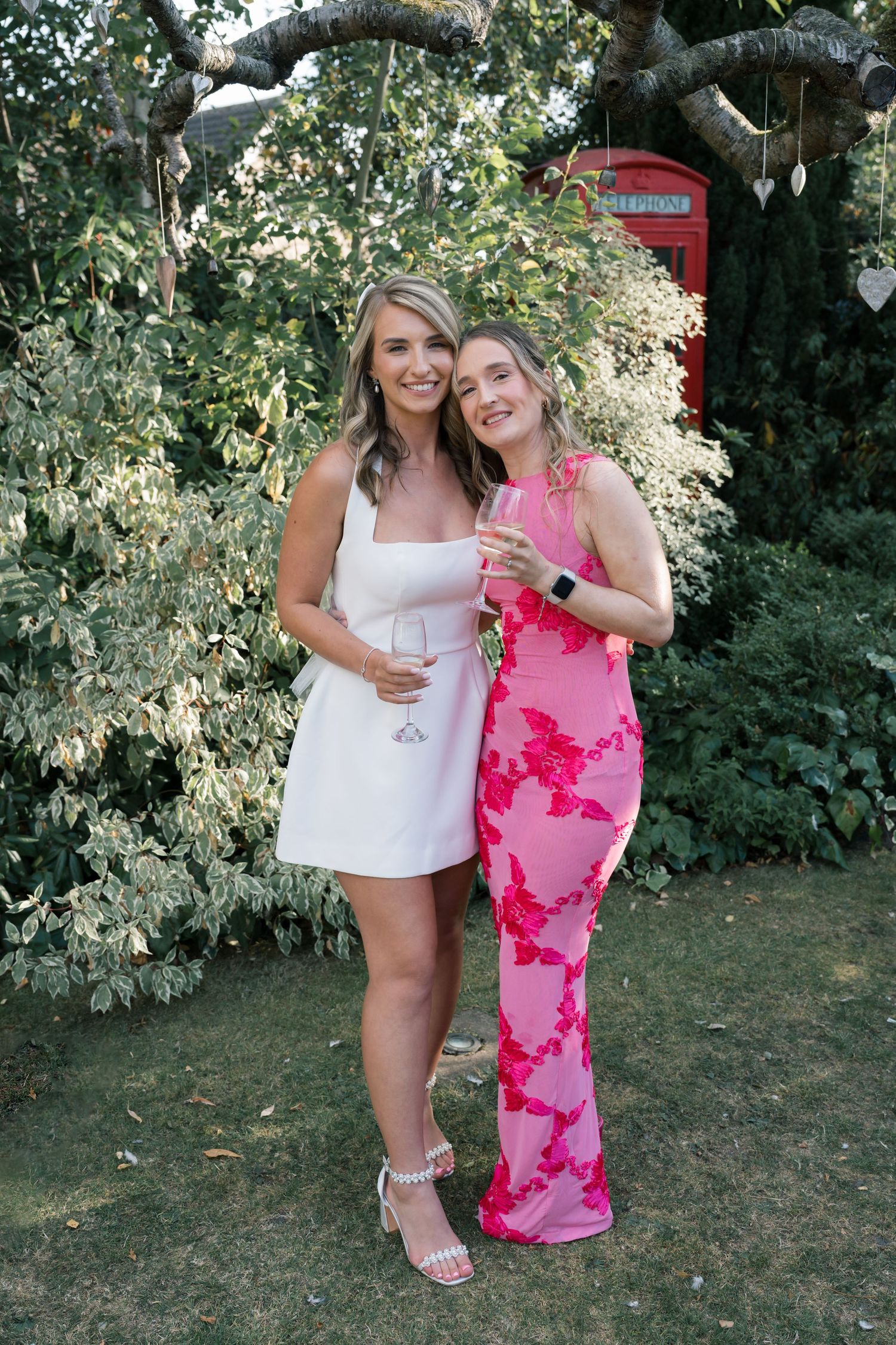 Two friends in summer outfits pose together outdoors in a garden setting with greenery in the background.