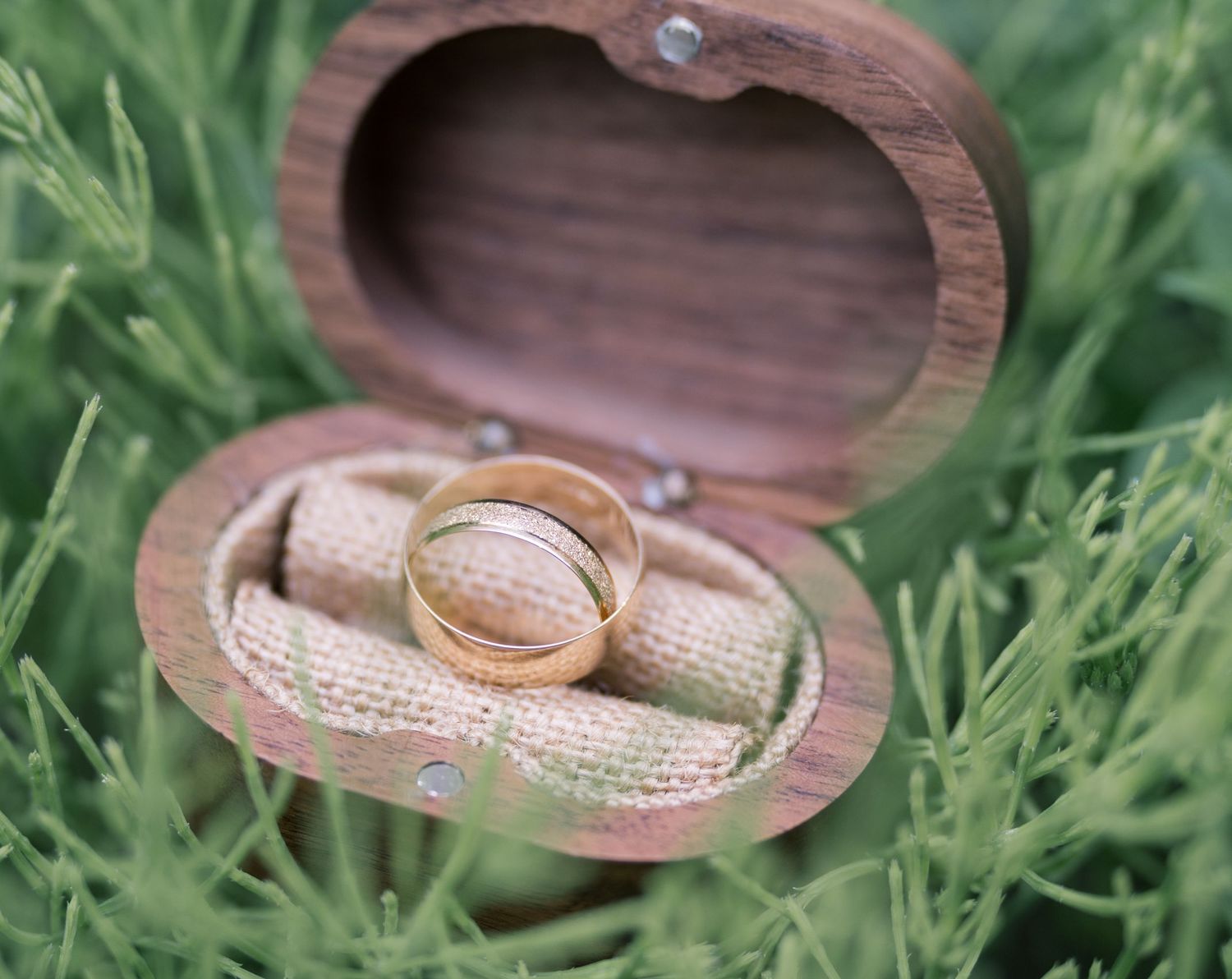 Two gold wedding rings rest inside a small wooden heart-shaped box nestled in green grass.