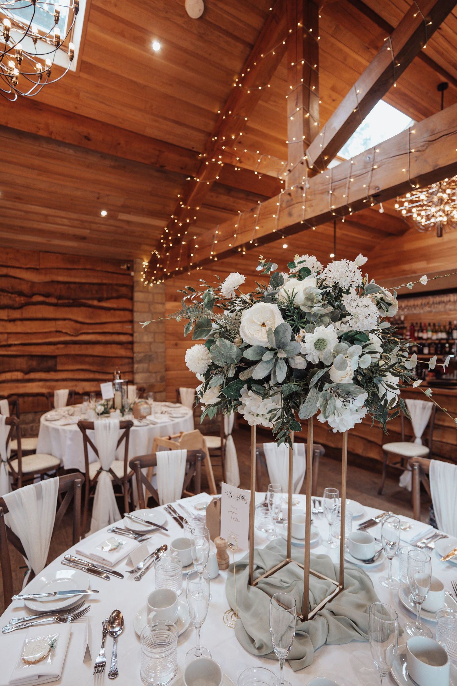 Elegant wedding reception table with tall white floral centerpiece and fairy lights in wooden venue.