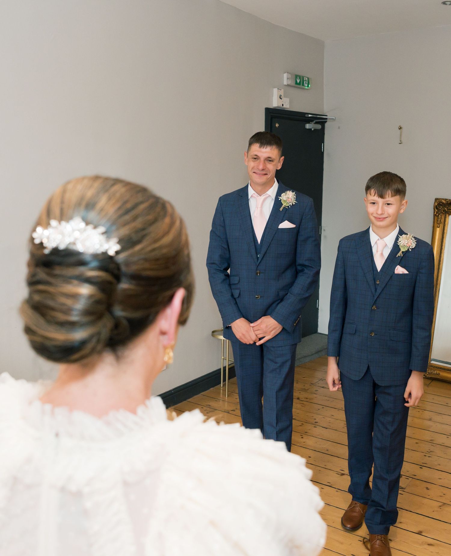 Wedding party members in formal attire stand in an indoor venue with wooden flooring.
