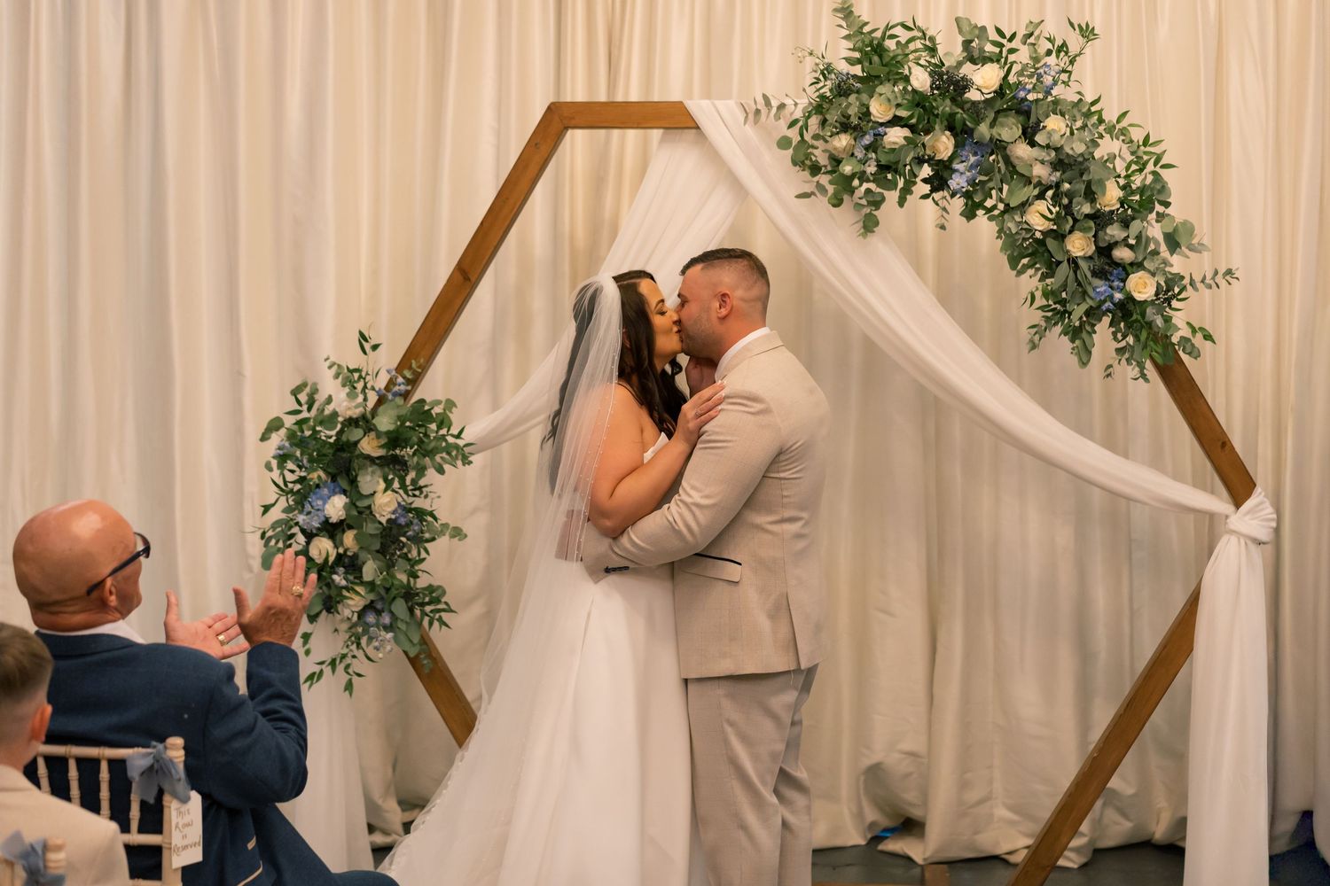 Wedding ceremony setup with wooden arch, white drapes, and greenery floral arrangements.
