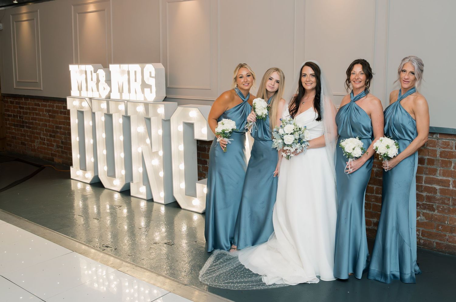 Wedding party in matching teal blue bridesmaid dresses pose with bride in white gown next to illuminated marquee letters.
