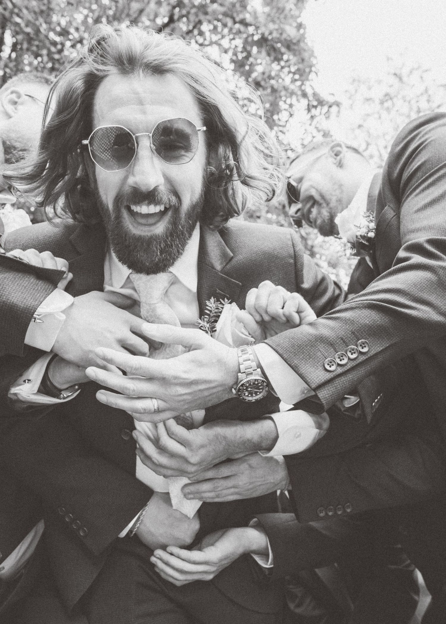 Groomsmen in dark suits playfully pose together outdoors at a wedding celebration.