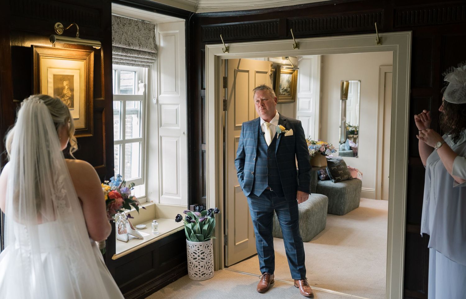 Father of the bride reacts emotionally seeing bride in white wedding dress and veil during first look moment indoors.