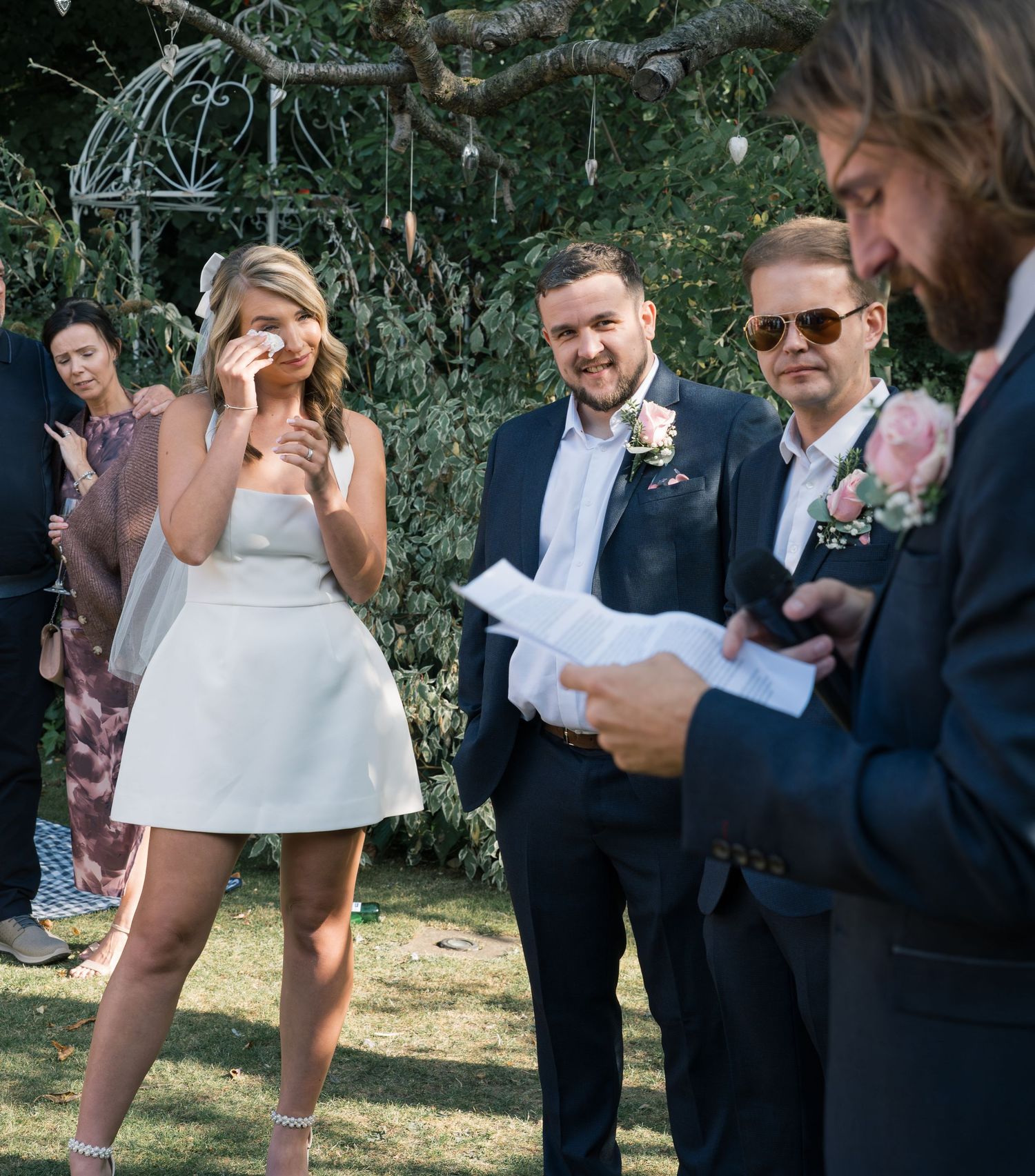 Wedding guests in formal attire gathered outdoors under trees for celebration.