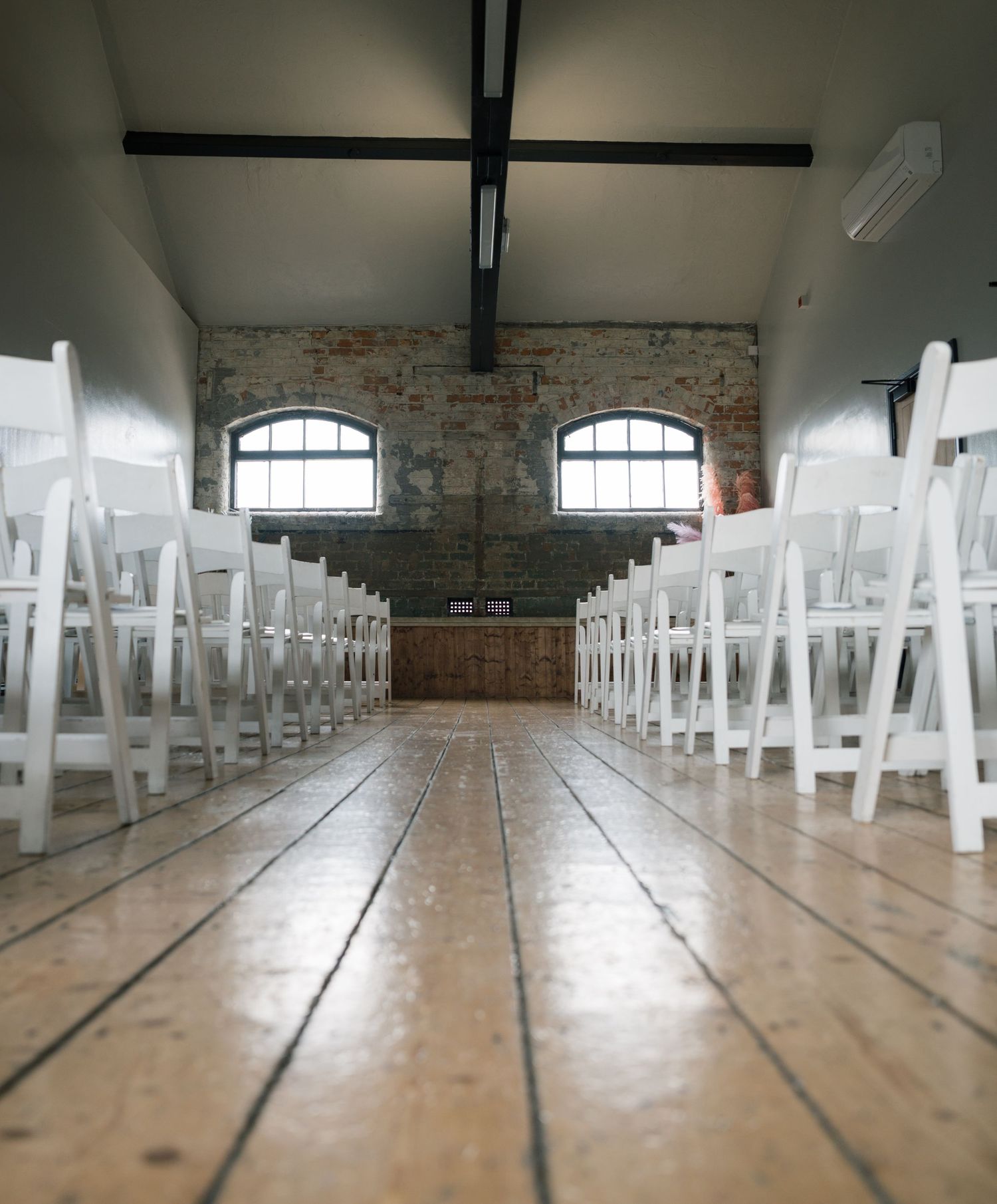 Empty wedding ceremony venue with rows of white chairs on wooden floors and exposed brick walls.