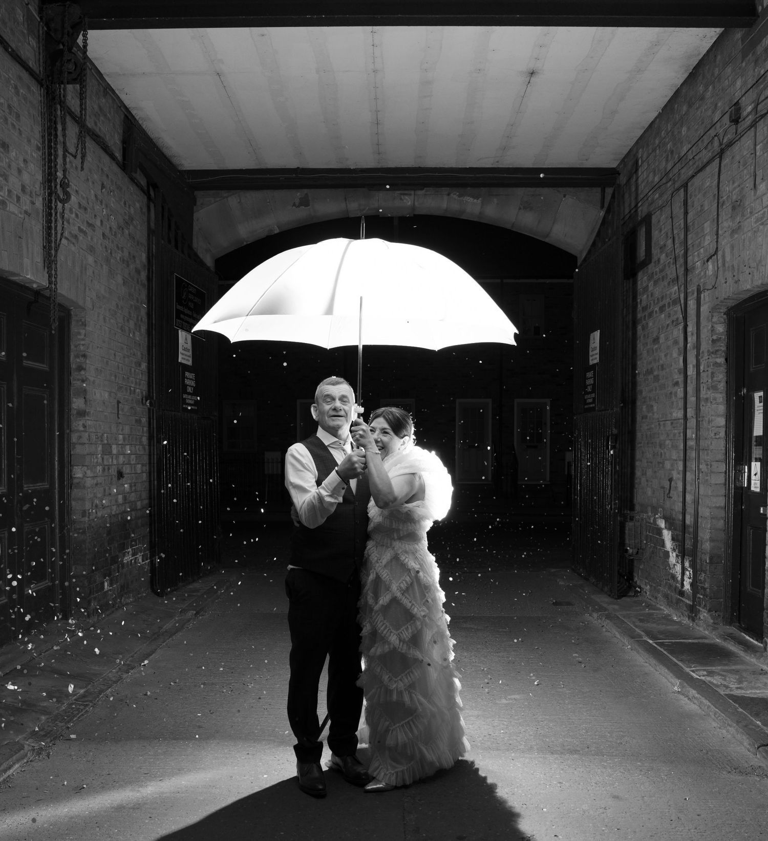 Black and white photo of a couple under an umbrella in a brick corridor.