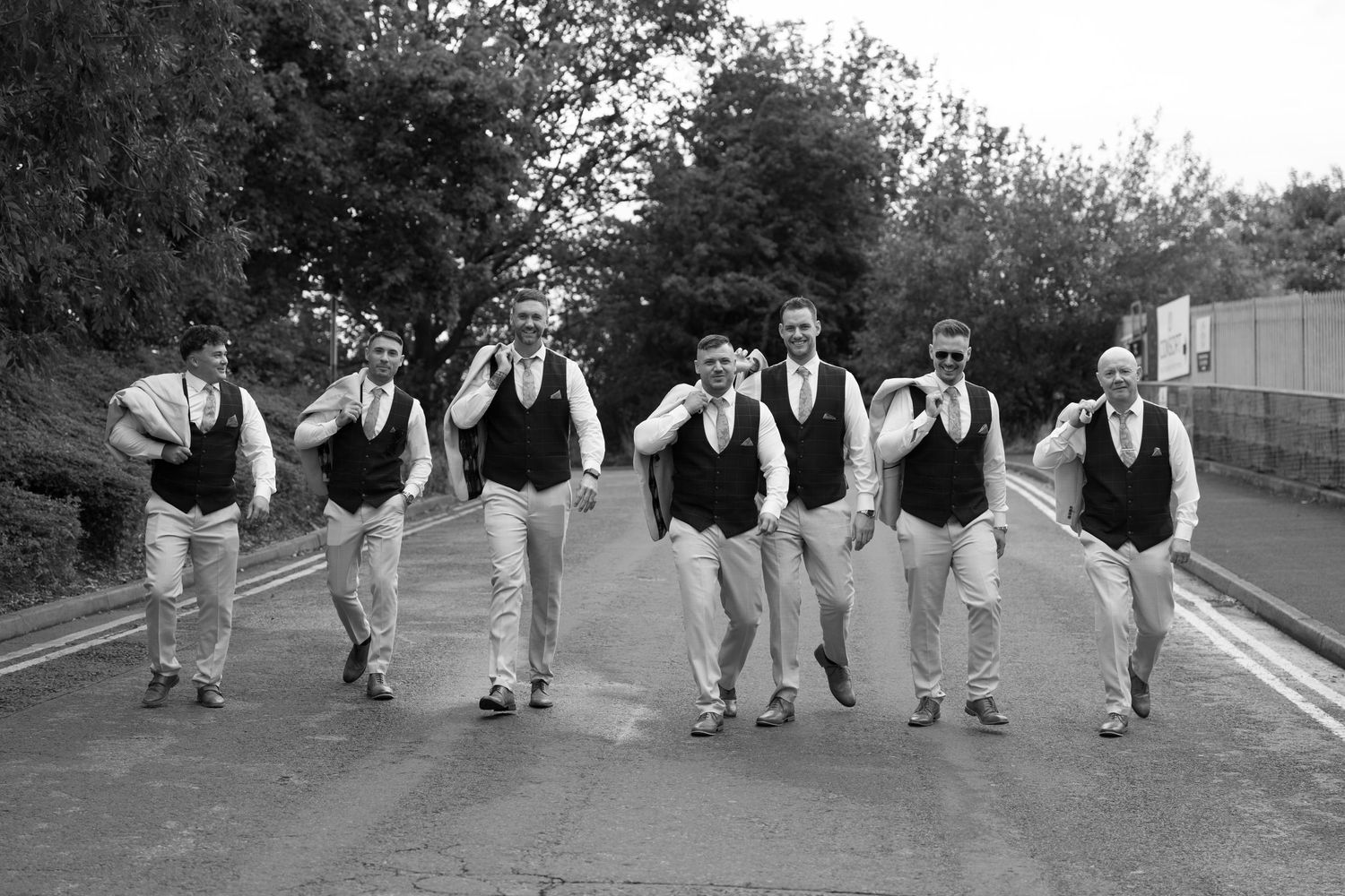 A group of groomsmen in matching black vests and white shirts walk together on a road in a black and white photo.