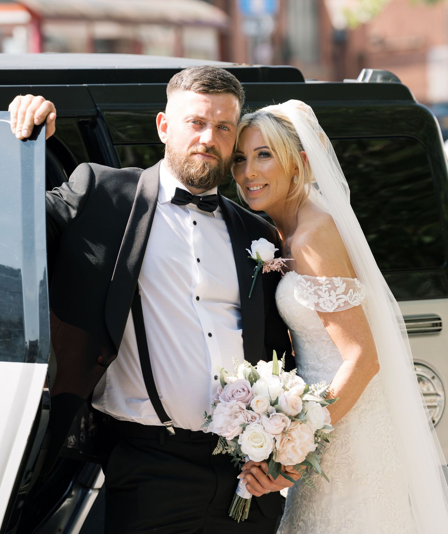 Bride and groom posing by a white limousine, bride holding a pastel bouquet in a lace off-shoulder gown.