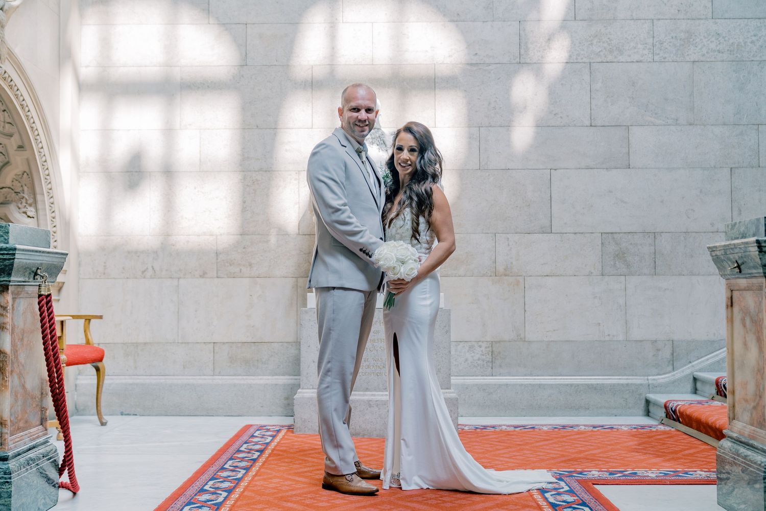 Wedding couple in light colored attire poses in front of white stone wall with arched window shadows.