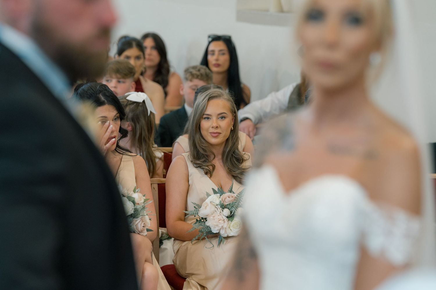 Emotional bridesmaids in blush dresses watch a wedding ceremony, one wiping tears, with bride in white gown, getting married in Leeds.
