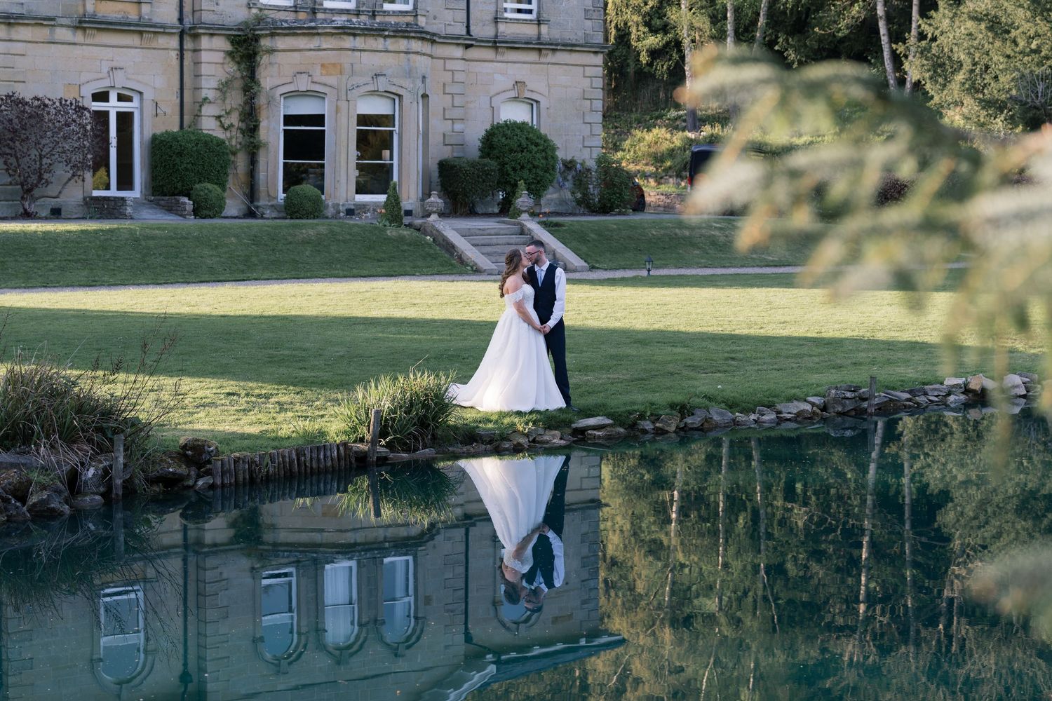 A wedding couple stands near a scenic golf course pond with lush green grass and trees in the background.