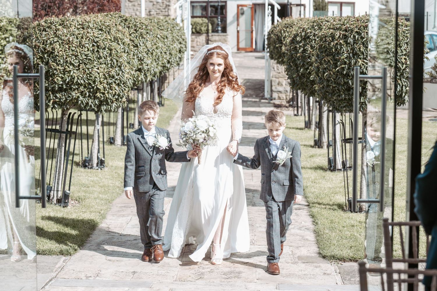 A wedding procession walking down a garden path lined with manicured hedges.