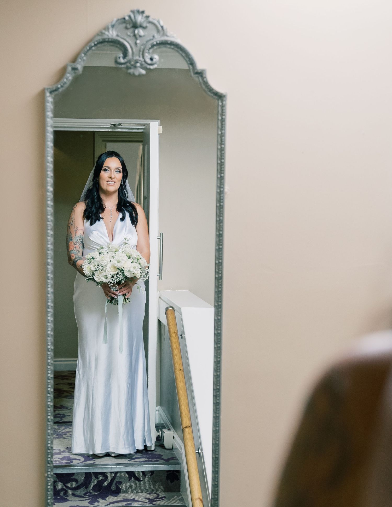 Bride in a white wedding dress stands in a doorway framed by an ornate mirror during a wedding preparation, at The Bridge Hotel & SPA, Selby