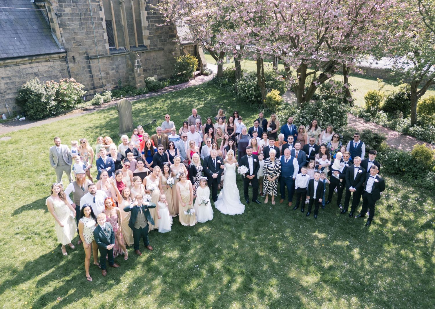 Aerial view of a large wedding party gathered on a green lawn beside a stone church with pink cherry blossom trees.