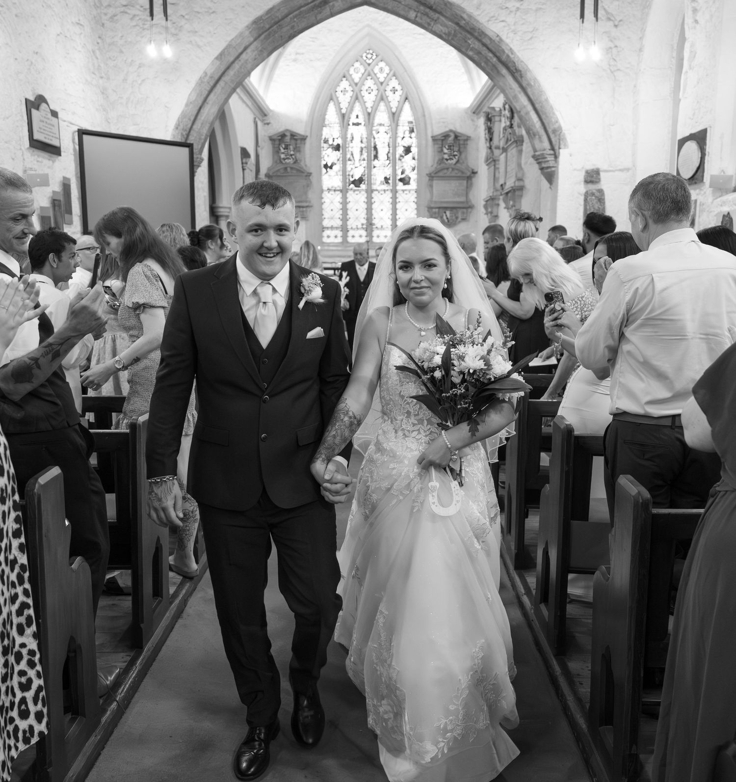 Black and white photo of newlyweds walking down church aisle after ceremony as guests look on in Gothic church interior.