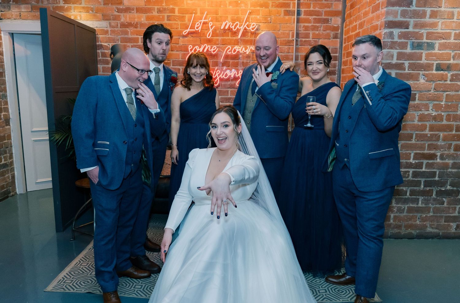Wedding party laughing together while posing against a rustic brick wall during an elegant indoor celebration.