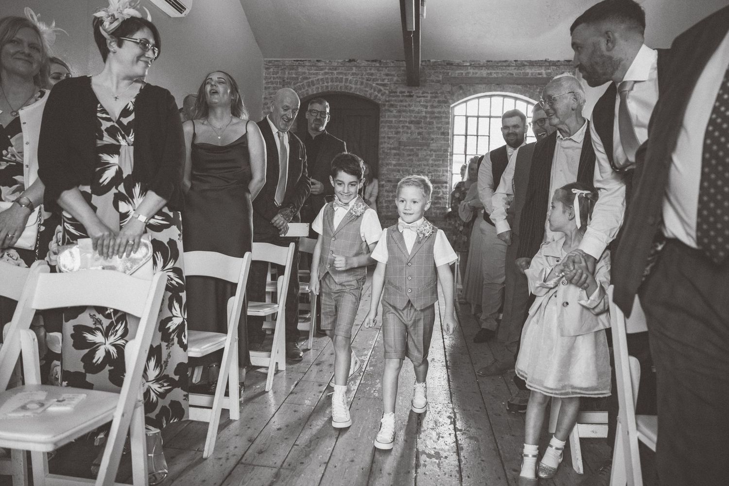 Black and white photo of wedding guests walking down aisle in rustic indoor venue.