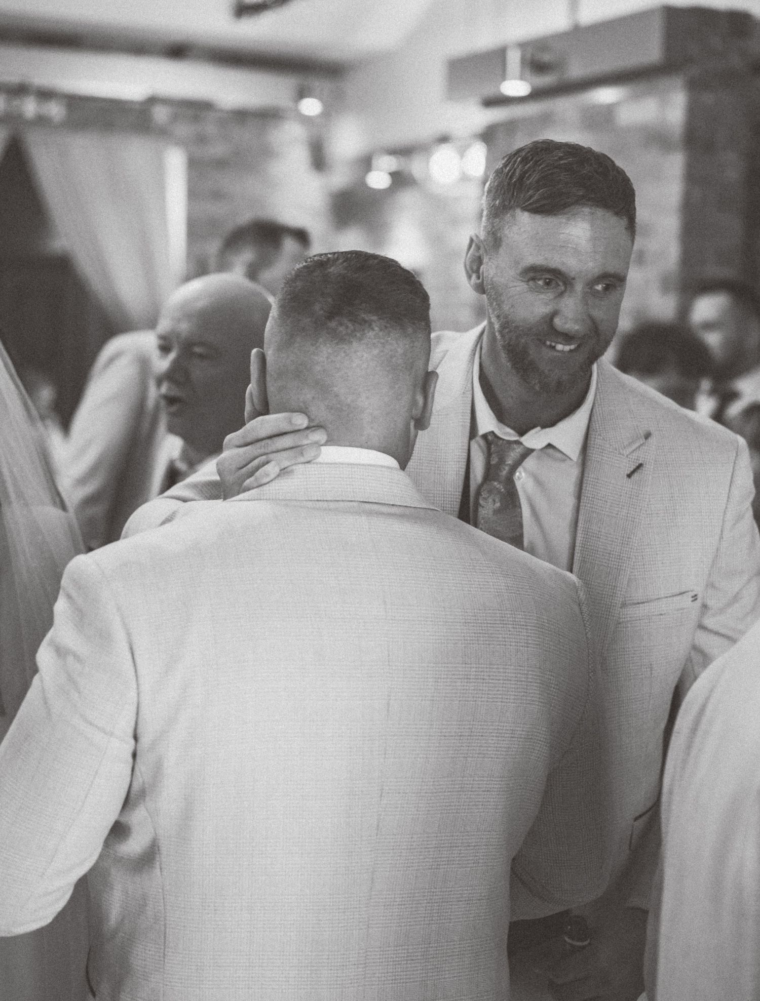 Two people in light colored suits sharing a moment at an indoor wedding reception.