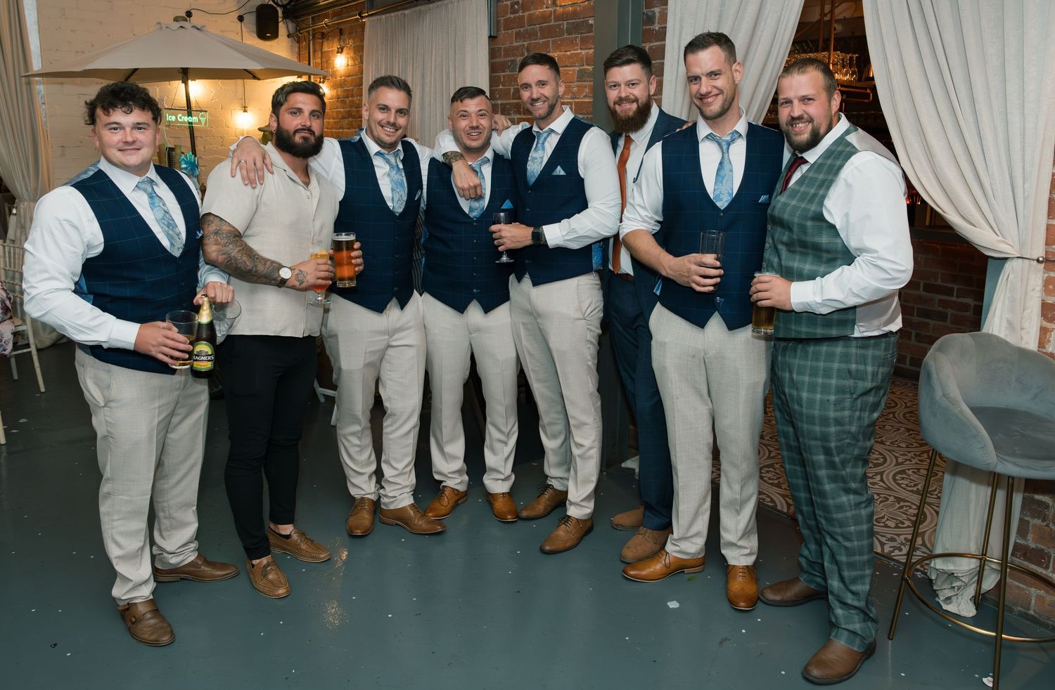 Groomsmen in matching vests and light pants pose together at a wedding reception with exposed brick walls.