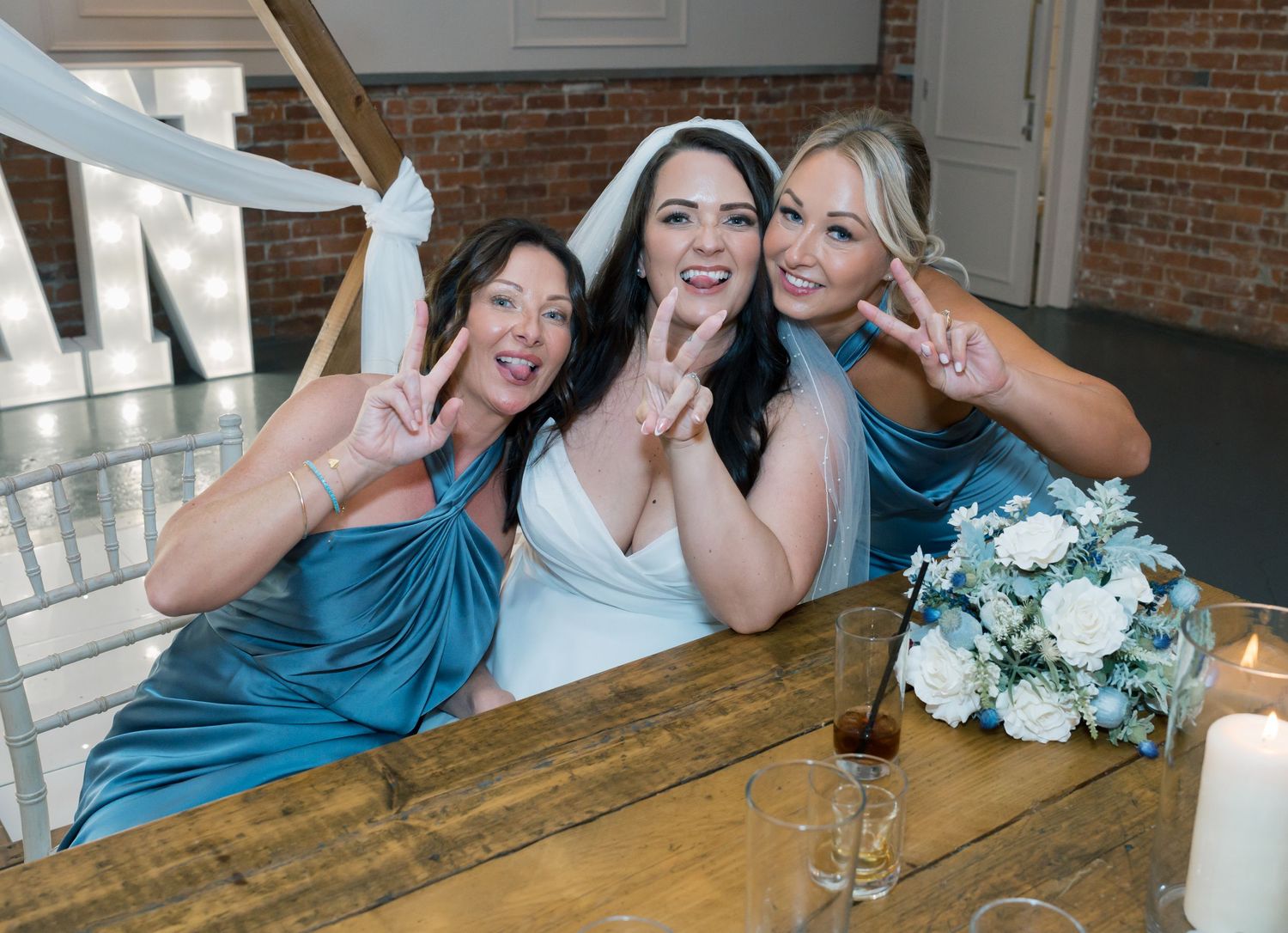 A group of wedding party members laugh and pose together at a rustic wooden table with a white floral bouquet.
