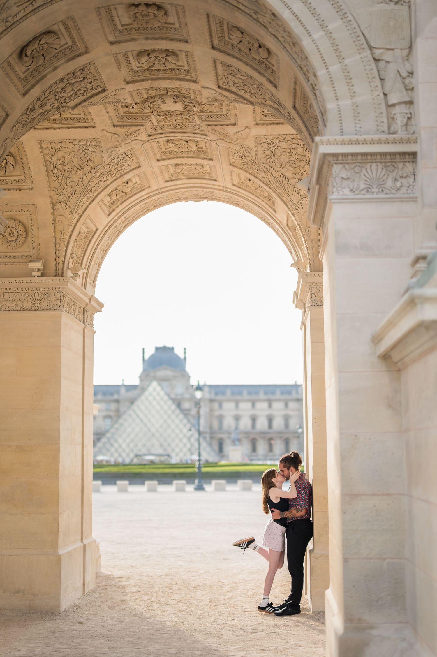Couple kissing under ornate arch at the Louvre Museum with glass pyramid in background, Paris.
