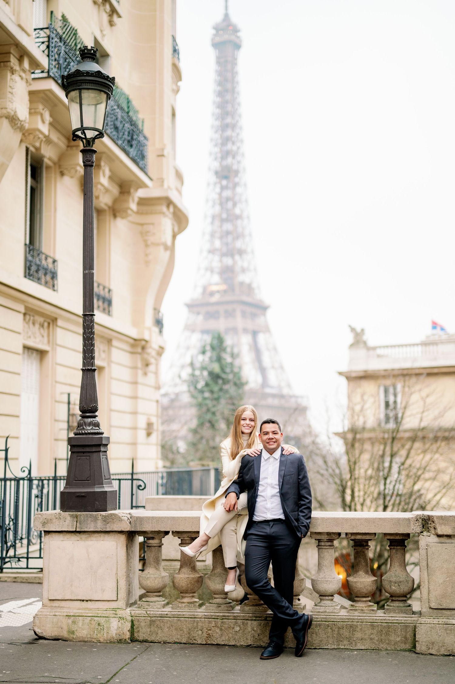 Stylish pair poses on ornate stone balustrade with Eiffel Tower in background.