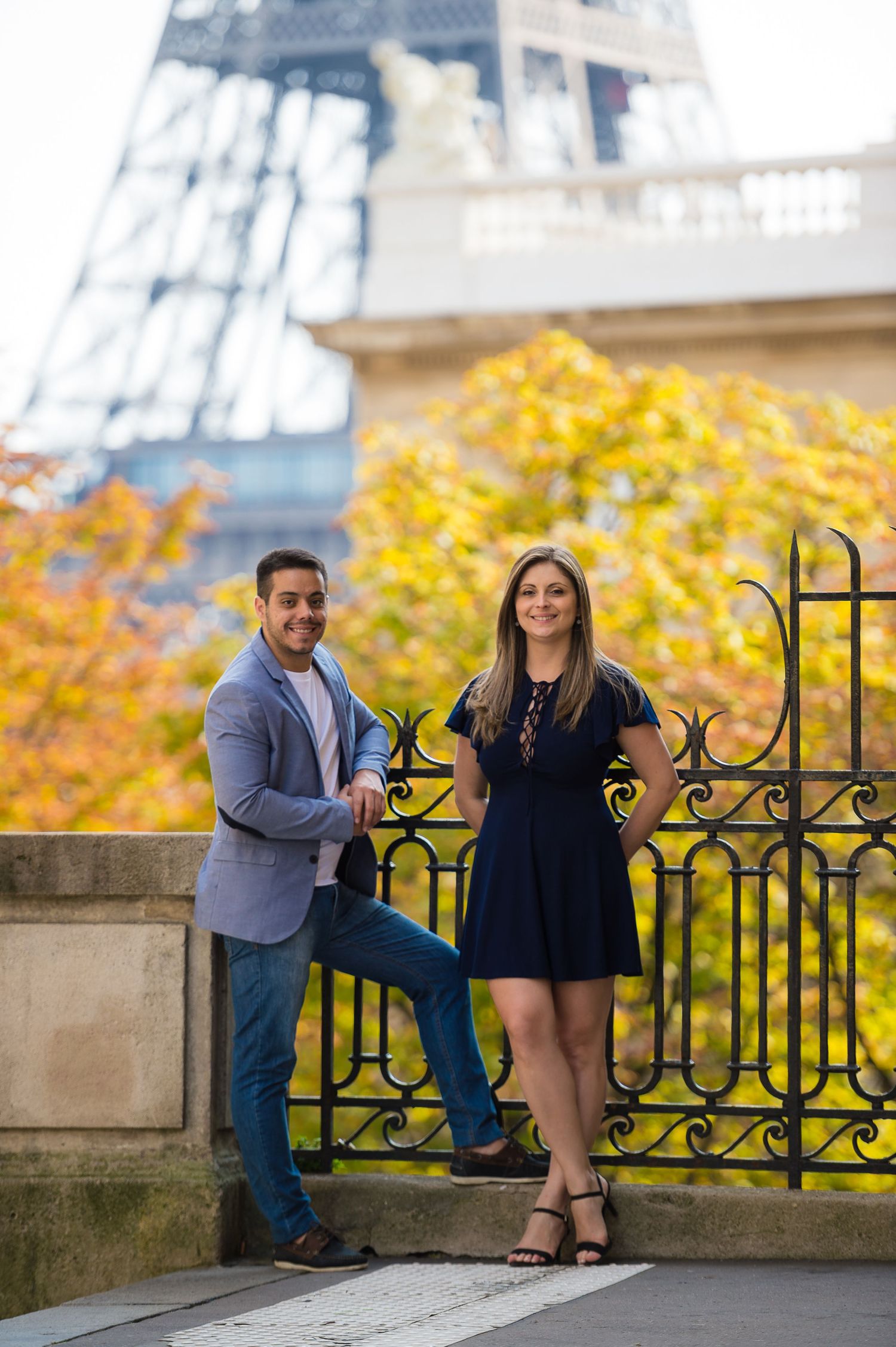 A couple poses by an ornate iron gate with autumn foliage and the Eiffel Tower visible in the background.