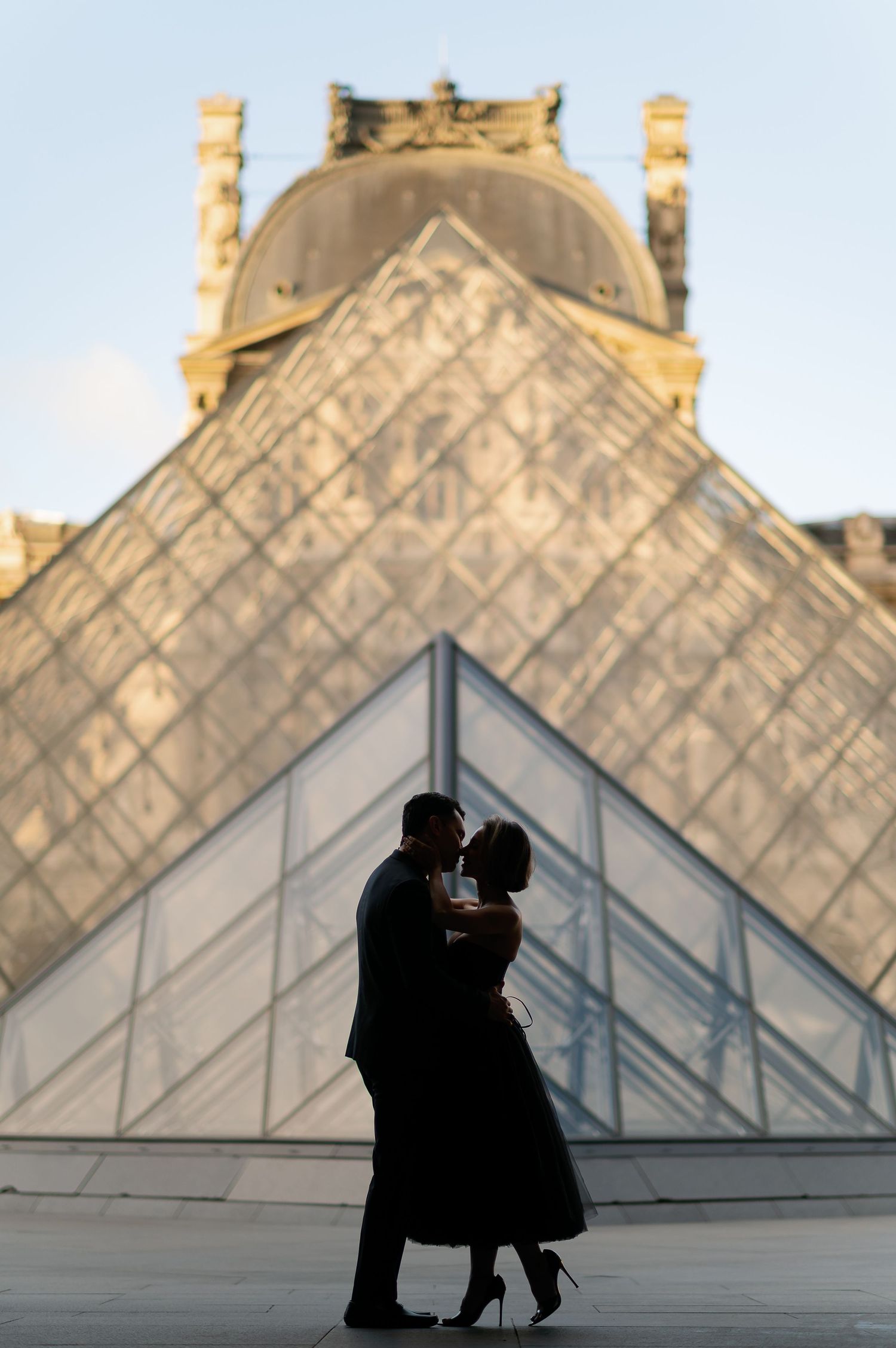 Couple silhouetted kissing in front of the Louvre Pyramid at golden hour in Paris, France.