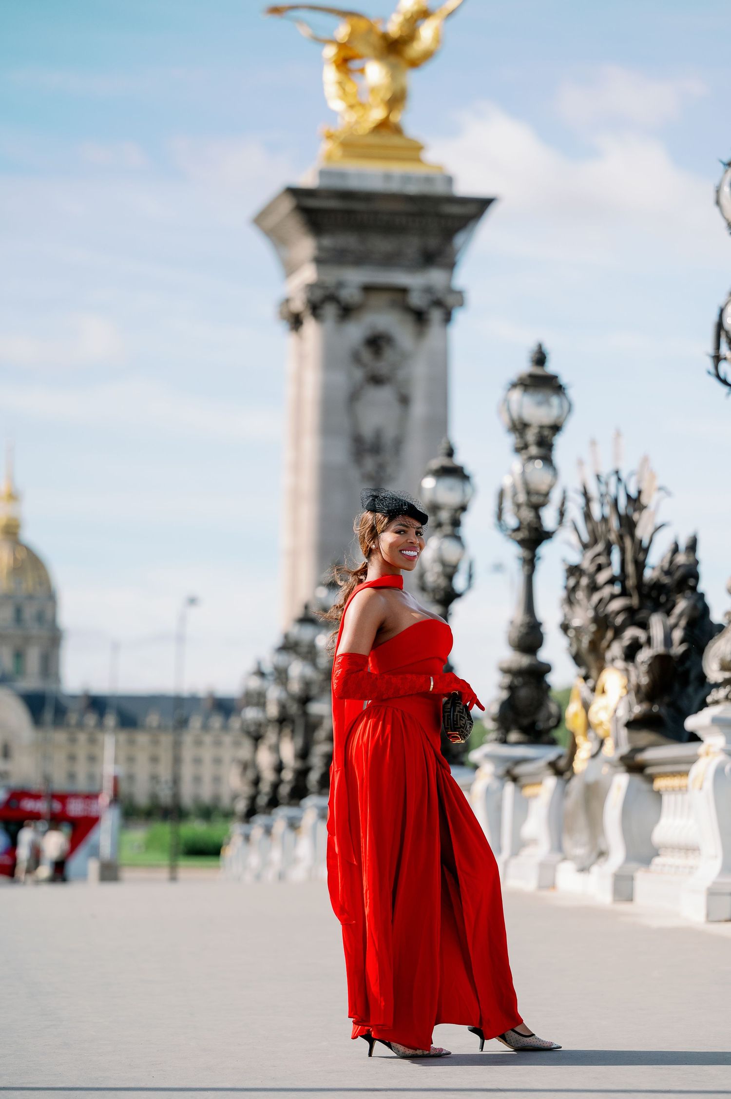 Woman in red gown poses on Pont Alexandre III bridge in Paris with golden statue and ornate lampposts.