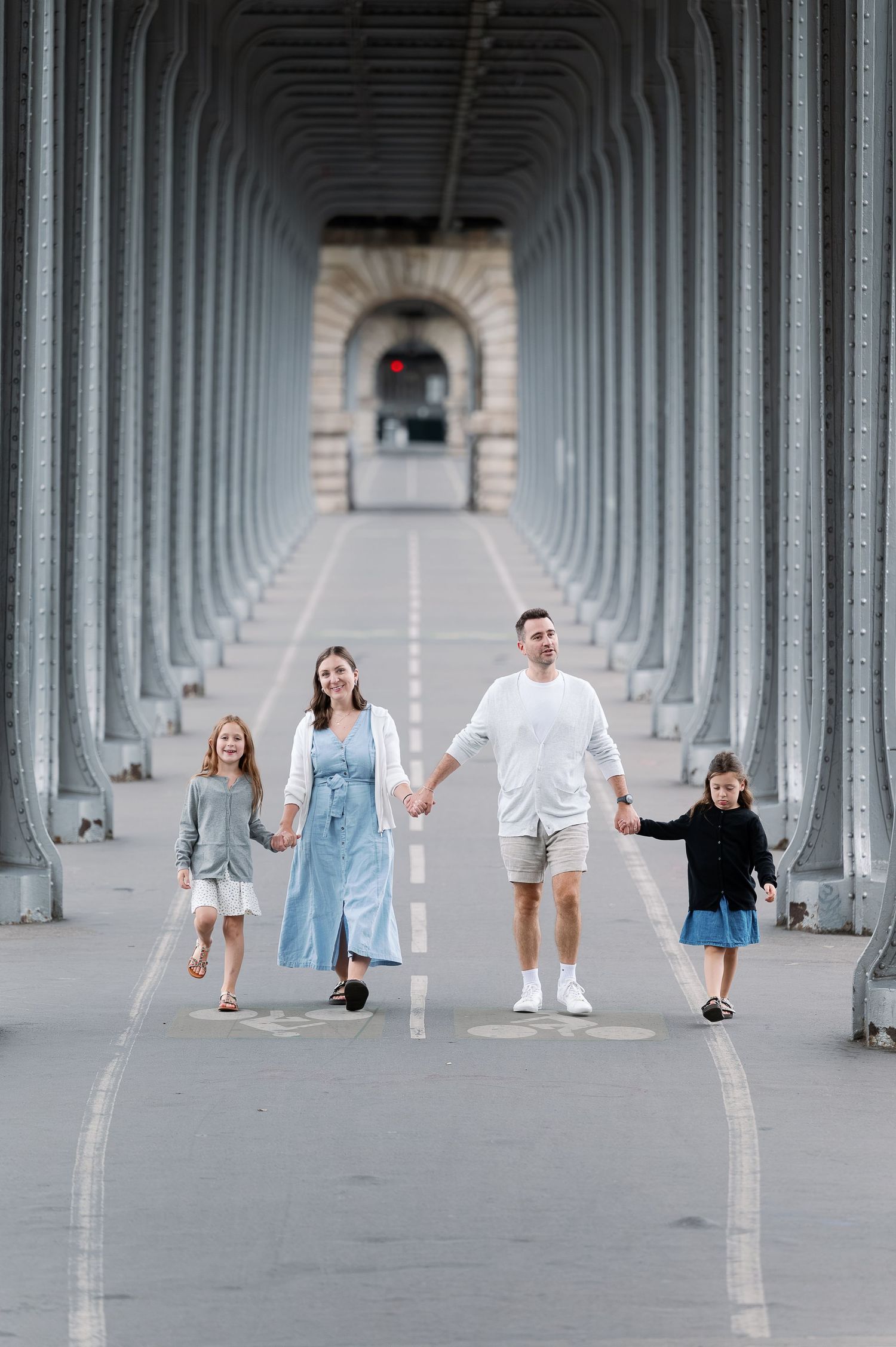 Family of four holding hands walking under a covered bridge with repeating arches in Paris.