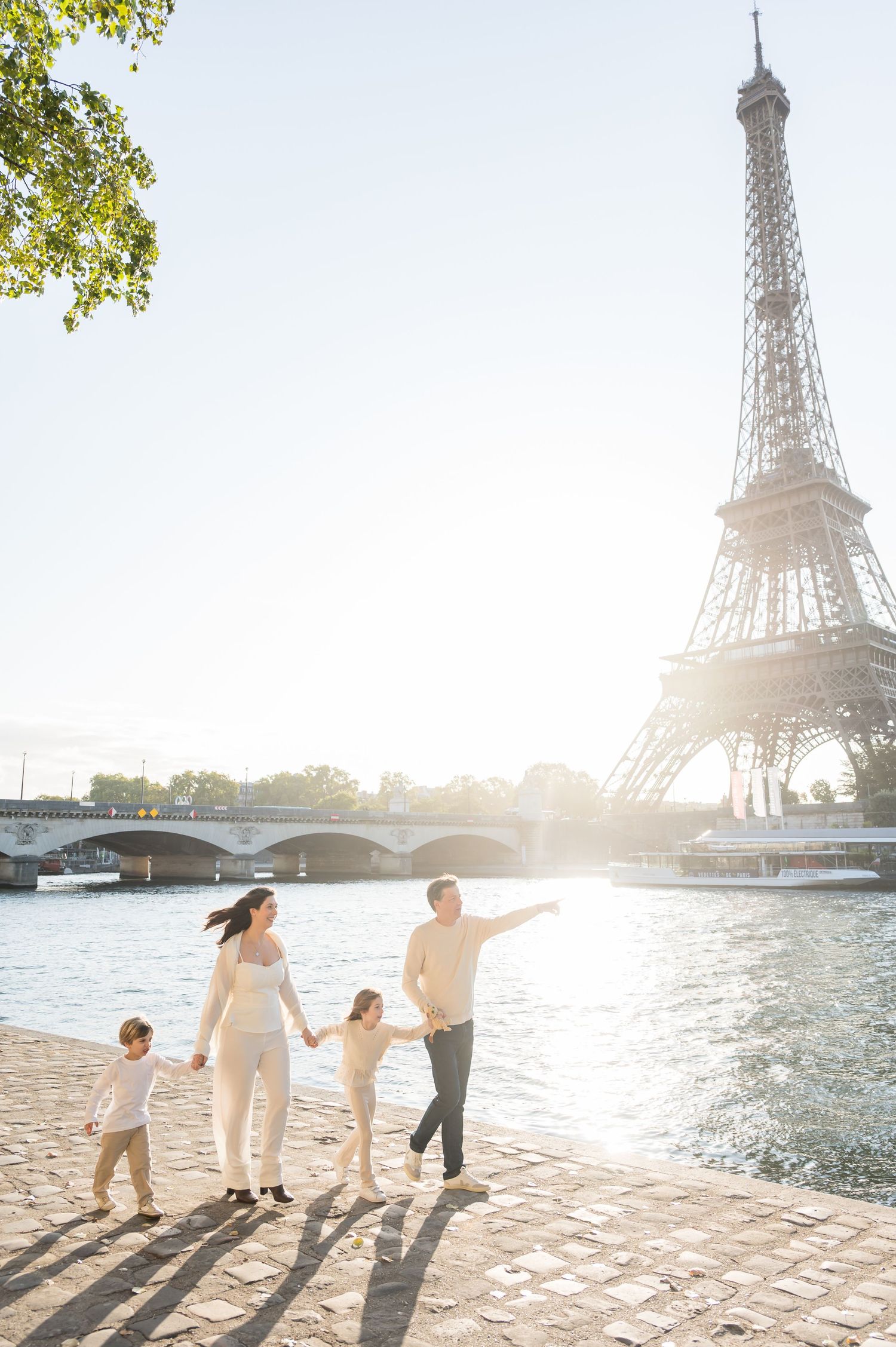Family walking hand in hand along the Seine River in Paris with Eiffel Tower and sunlight in background.