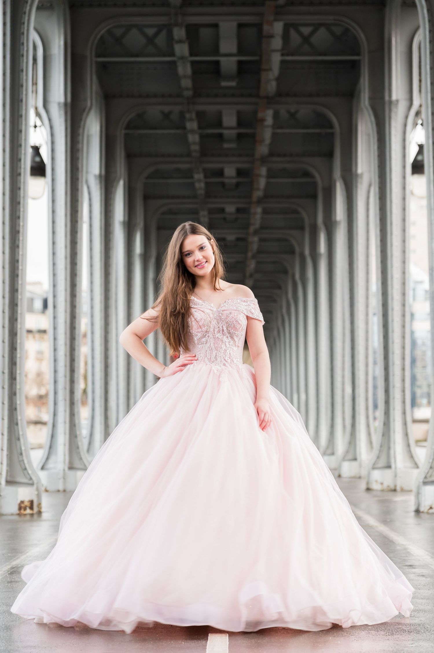 A person in a flowing pink ballgown poses elegantly under the arches of Bir-Hakeim Bridge in Paris.