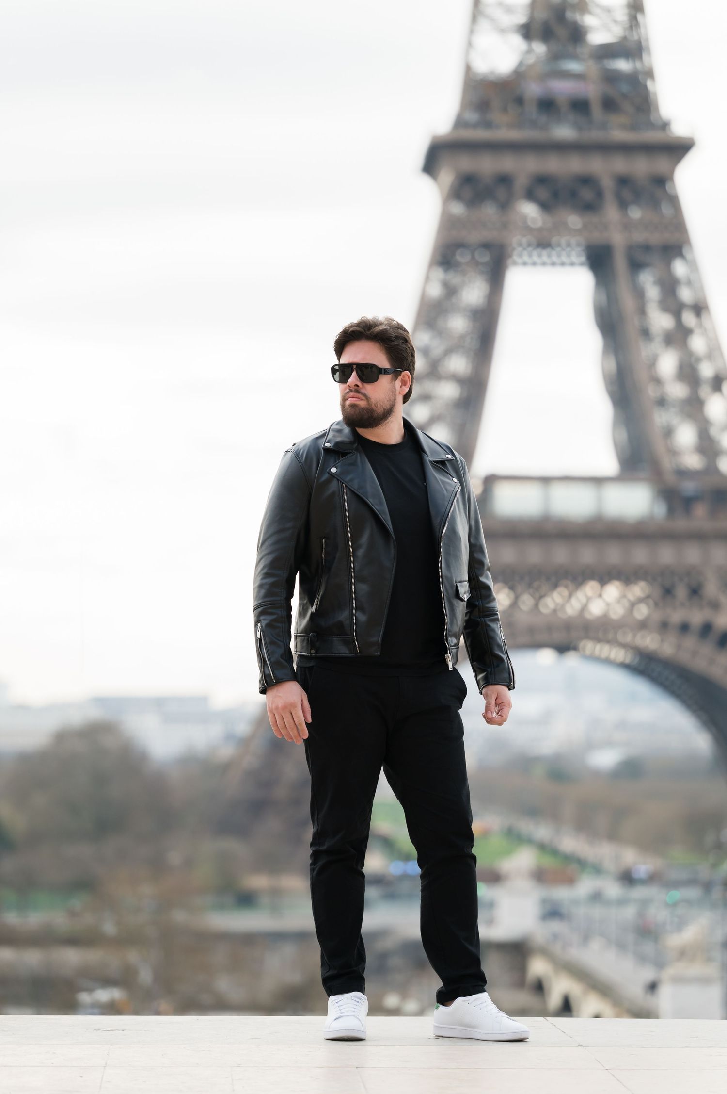 A bearded man in a black leather jacket and sunglasses poses confidently in front of the Eiffel Tower in Paris.