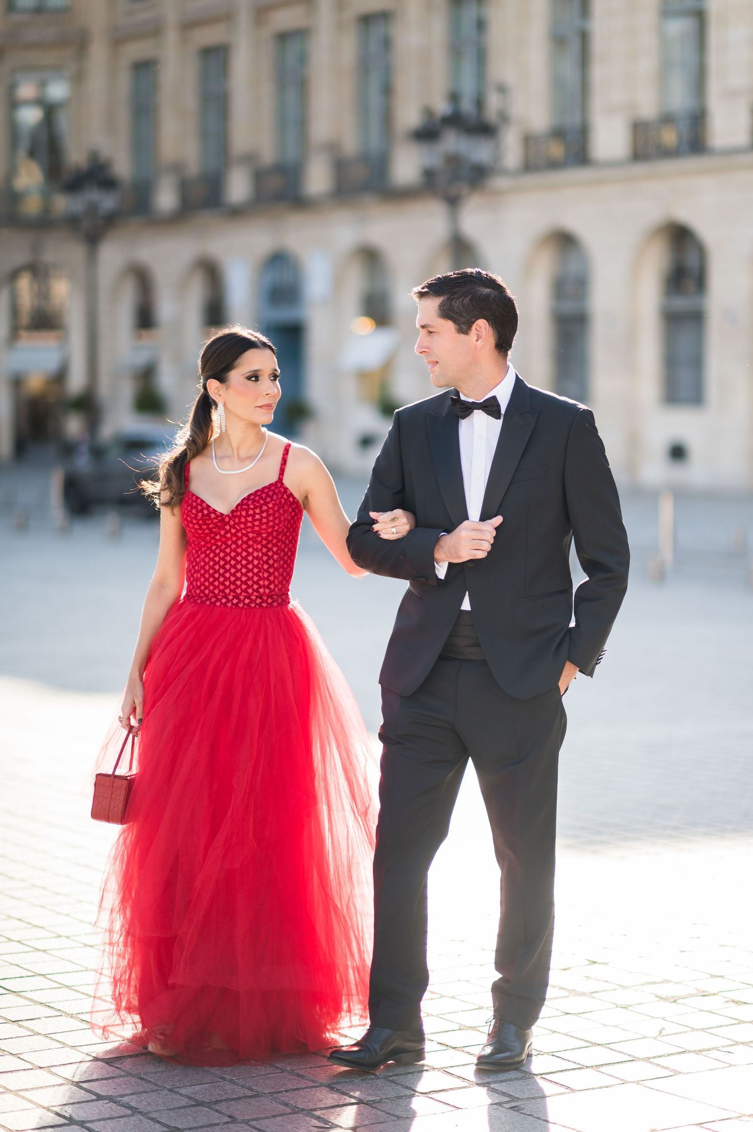 Elegant couple in formal attire, woman in red tulle gown and man in black tuxedo, walking in a sunlit plaza.