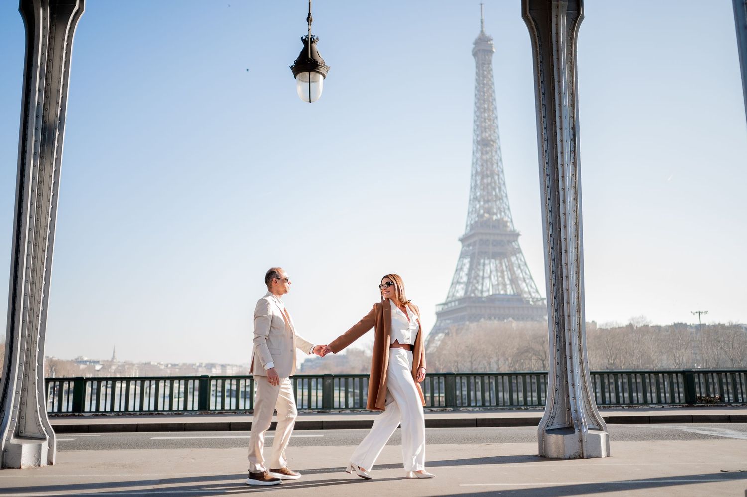 A couple in white outfits holds hands on a Parisian bridge with the Eiffel Tower in the background.