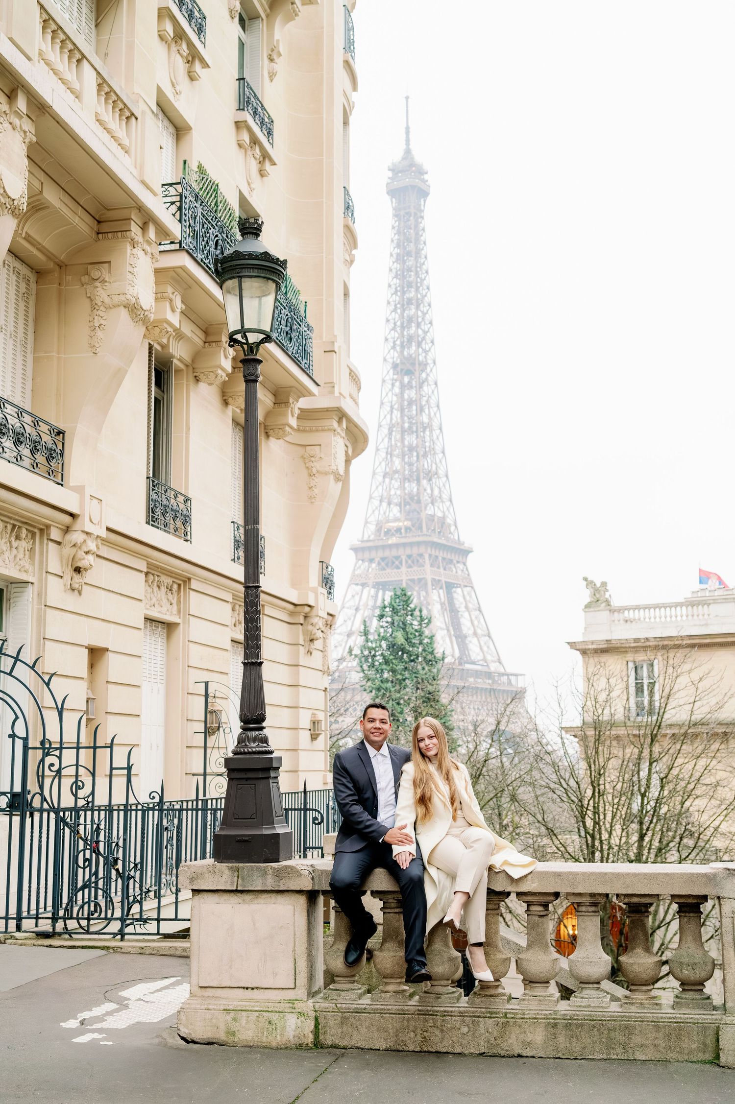 Couple sitting on stone balustrade against classic Parisian architecture with Eiffel Tower backdrop.