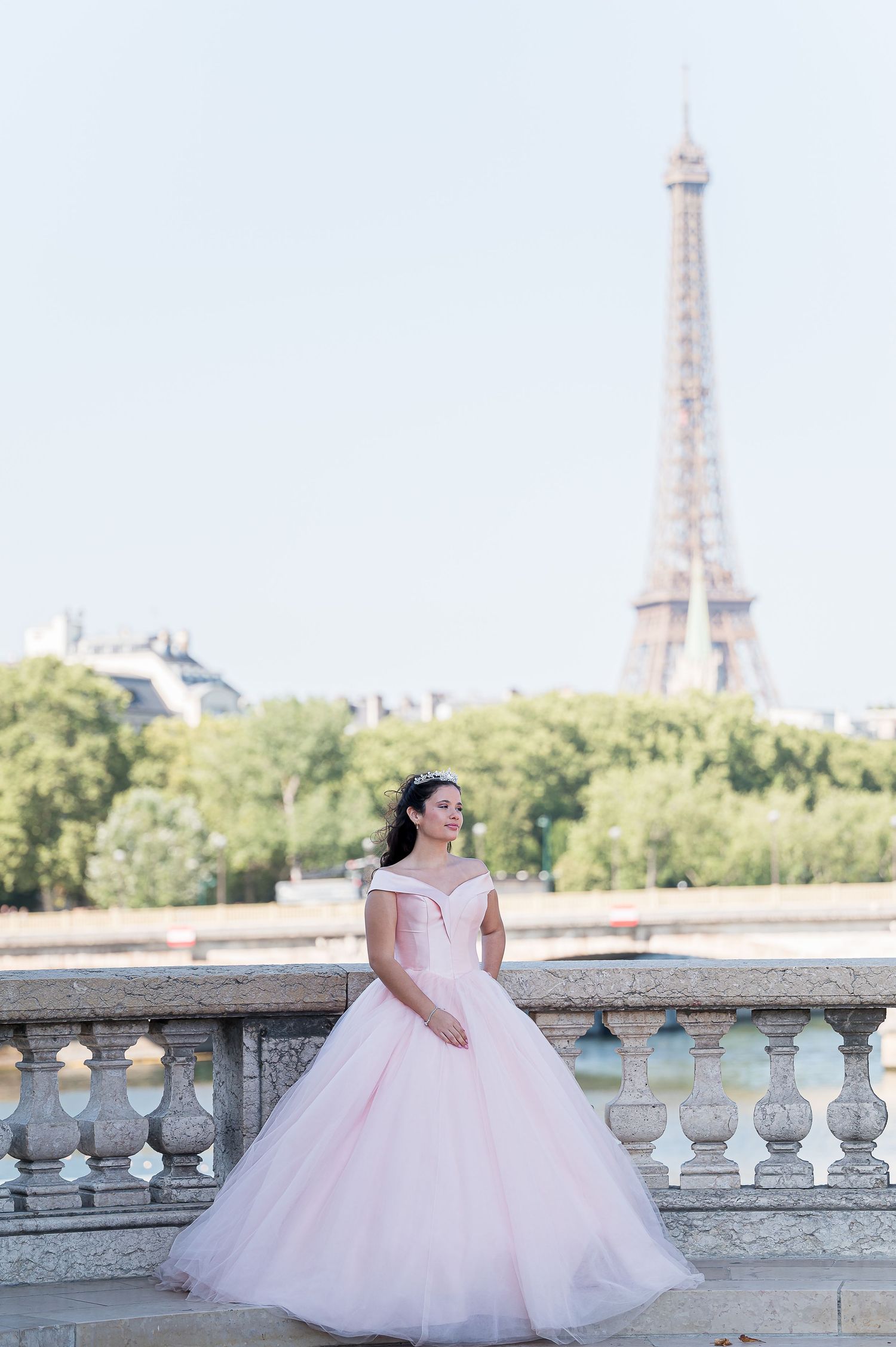 Woman in pink ballgown poses on a bridge with the Eiffel Tower in Paris behind her.