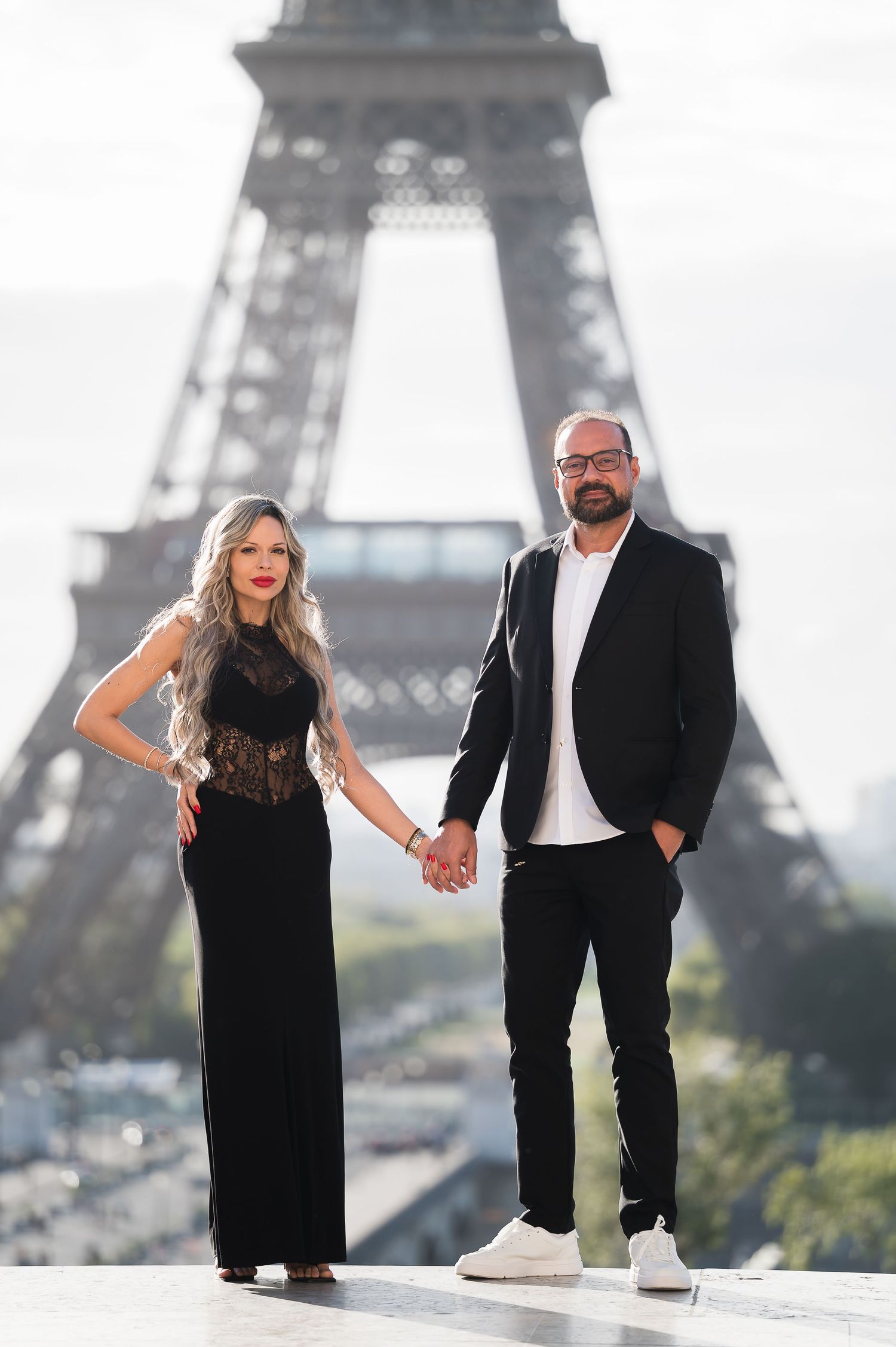 Couple holding hands in front of the Eiffel Tower in Paris, woman in black gown and man in black suit.